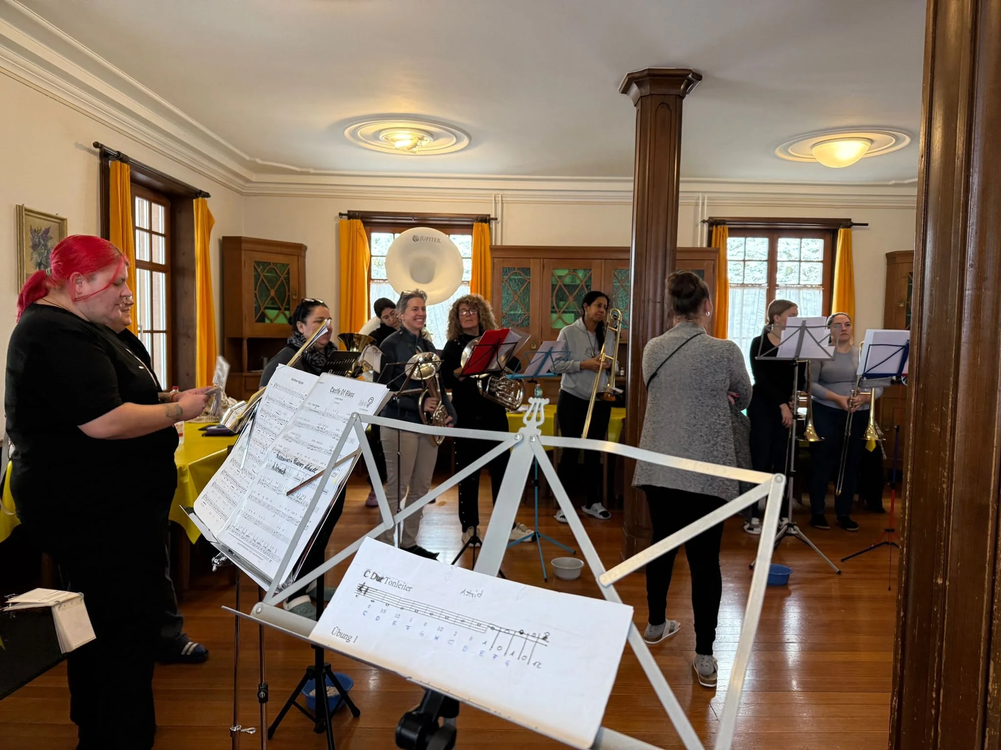 Musikerinnen bereiten sich auf ein Proben- oder Auftrittssituation in einem Raum mit Holzfußböden und Fenstern mit gelben Vorhängen vor.
