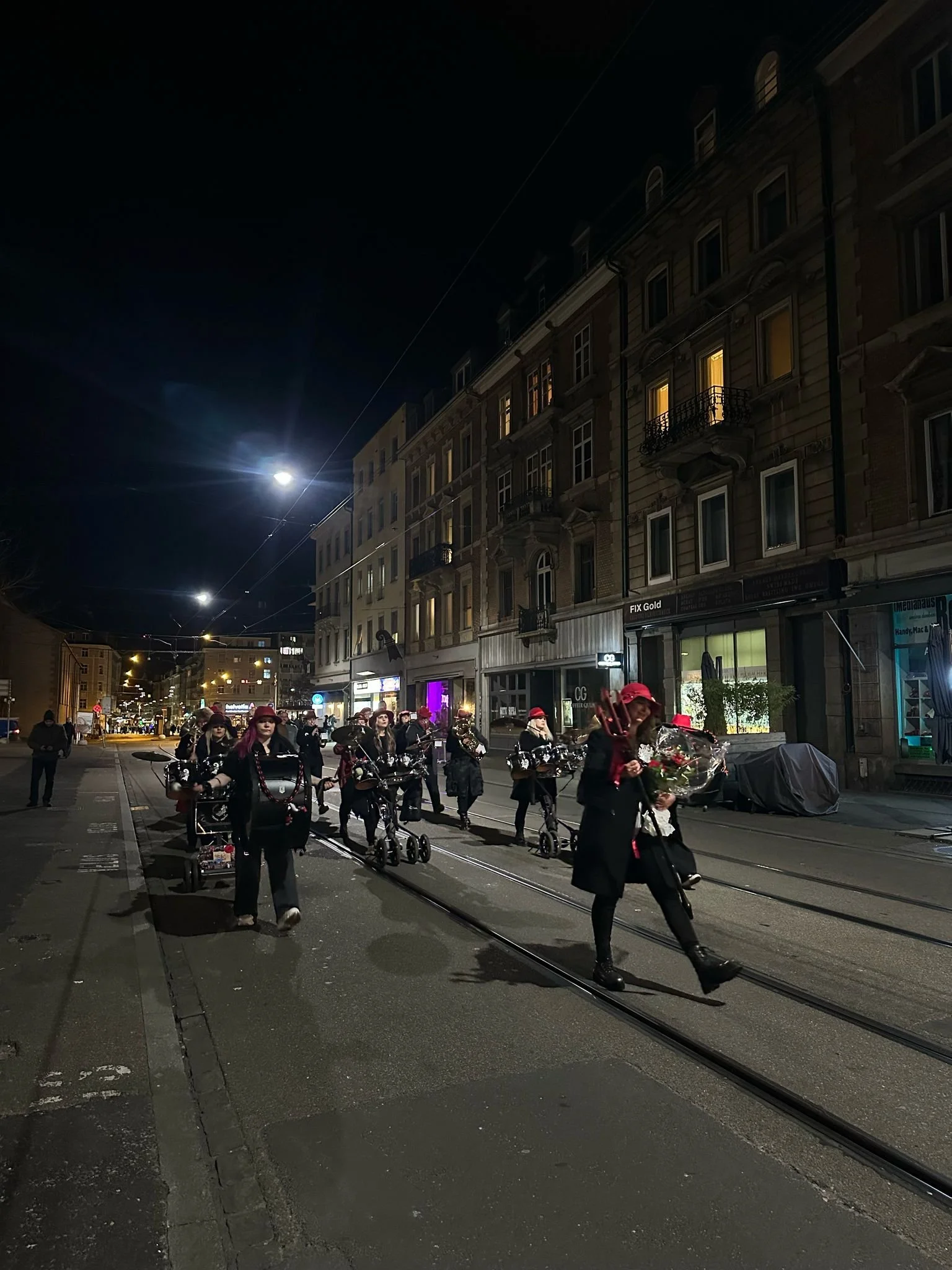 Gruppe von Menschen, die im Dunkeln auf Straßenbahngleisen in einer Stadt bei Nacht gehen, mit Gebäuden im Hintergrund.