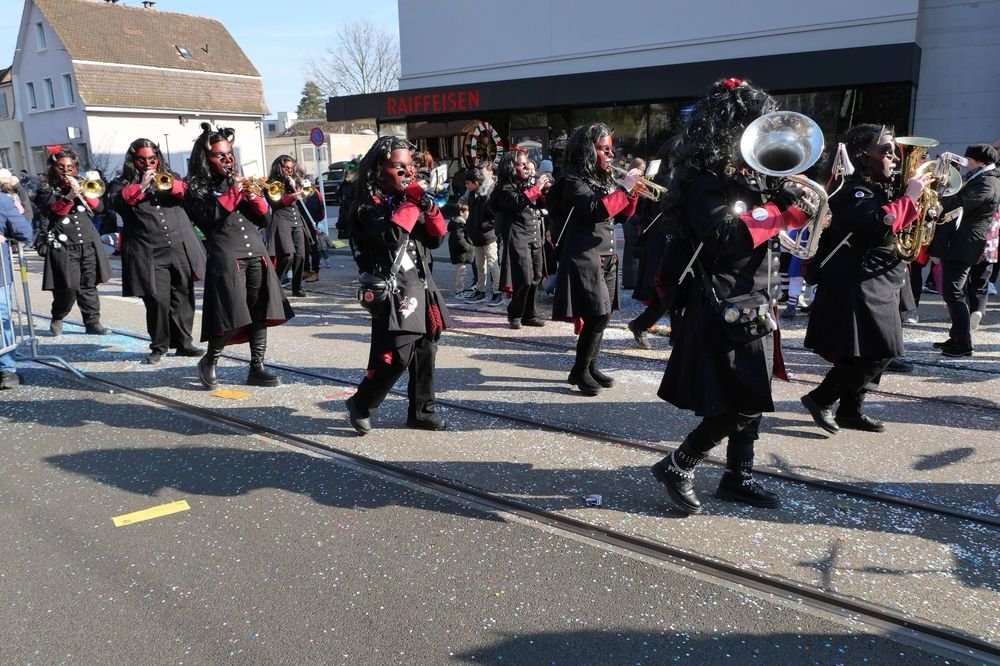 Eine Gruppe von Menschen in schwarzer Kleidung mit roten Akzenten, die auf einer Straße marschieren und Musikinstrumente spielen, während sie an einem Parade- oder Festumzug teilnehmen.