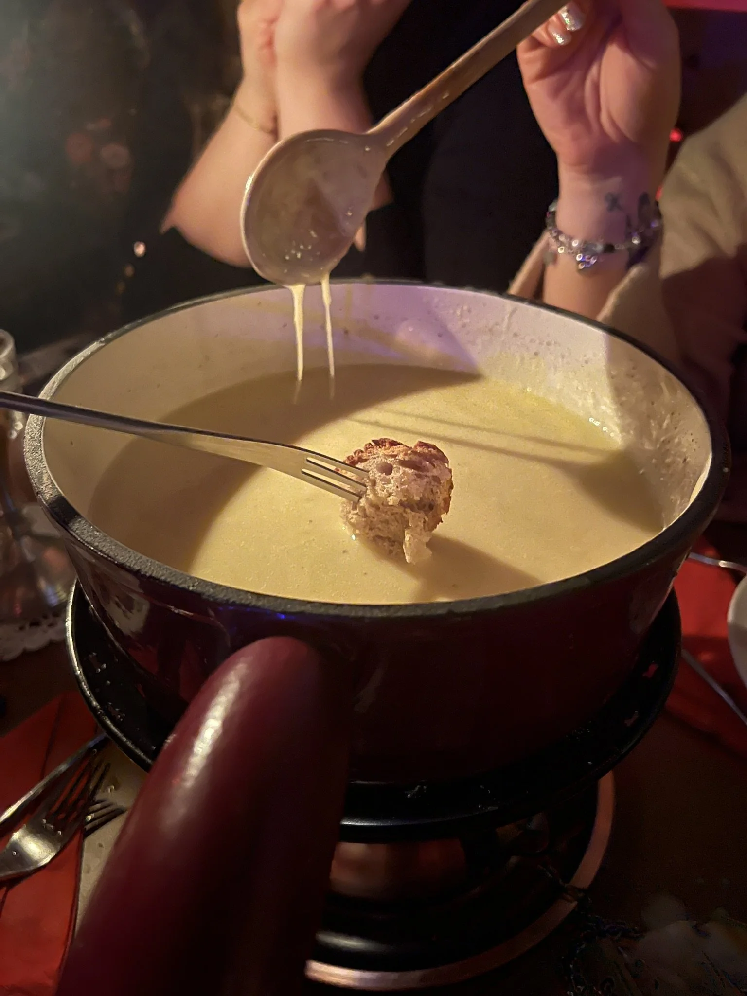 Person taucht einen Käsespieß in eine neue Käsesauce. Im Hintergrund trägt jemand eine Schildkappe und eine Armbanduhr.