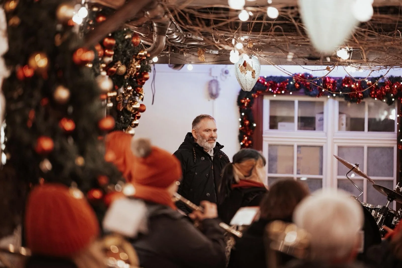 Menschen, die bei einer Weihnachtsfeier in einer festlich dekorierten Umgebung Musik machen, mit Weihnachtskugeln und Lichtergirlanden. Das Bild zeigt einen Mann, der im Hintergrund steht, und mehrere Personen, die ein Instrument spielen.