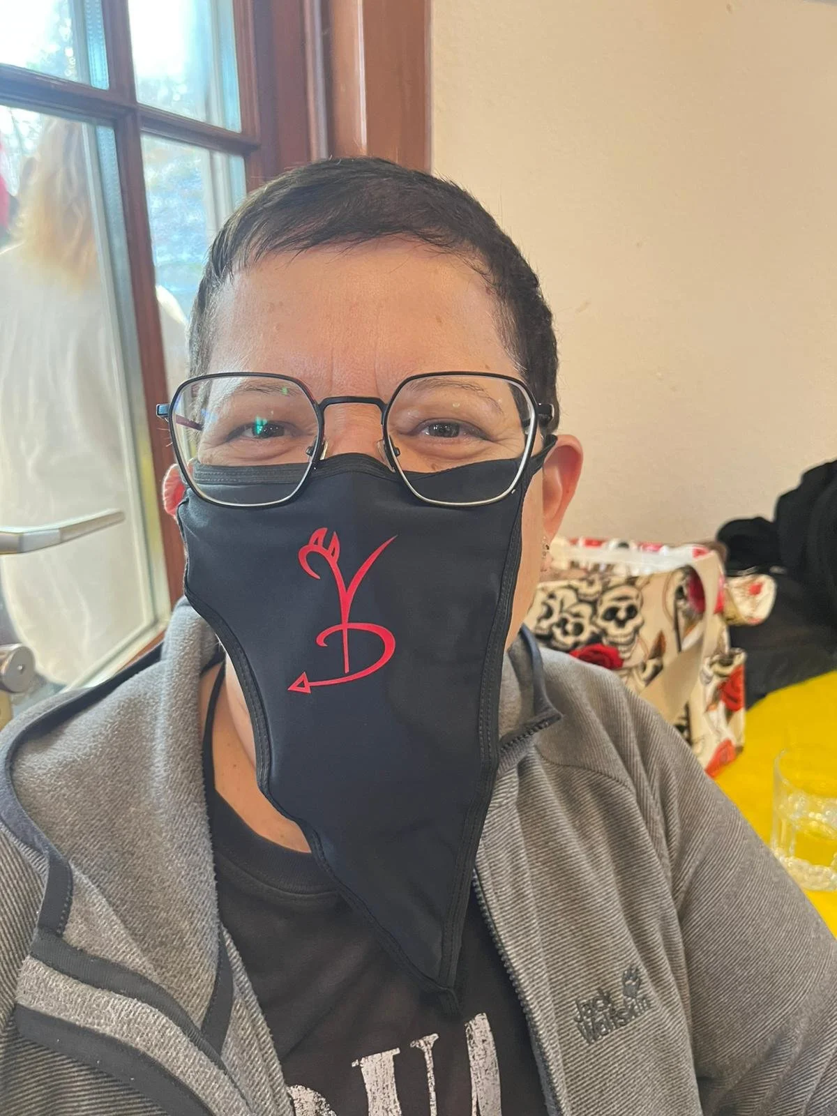 Ein Mann trägt eine schwarze Gesichtsmaske mit einem roten Pfeil- und Teufel-Logo, liest oder trägt eine Brille, sitzt an einem Tisch in einem Raum mit Holztüren oder Fenstern im Hintergrund.