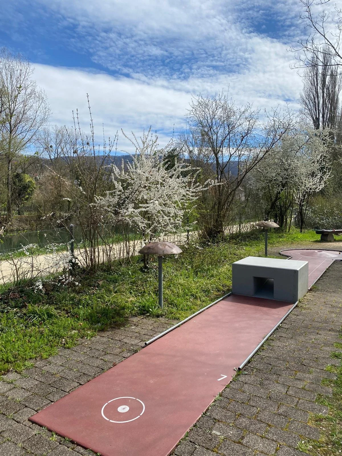 Minigolfanlage im Freien mit künstlichen Bahnen und Hindernissen, umgeben von blühenden Bäumen und einer Landschaft im Hintergrund, bei schönem Wetter.