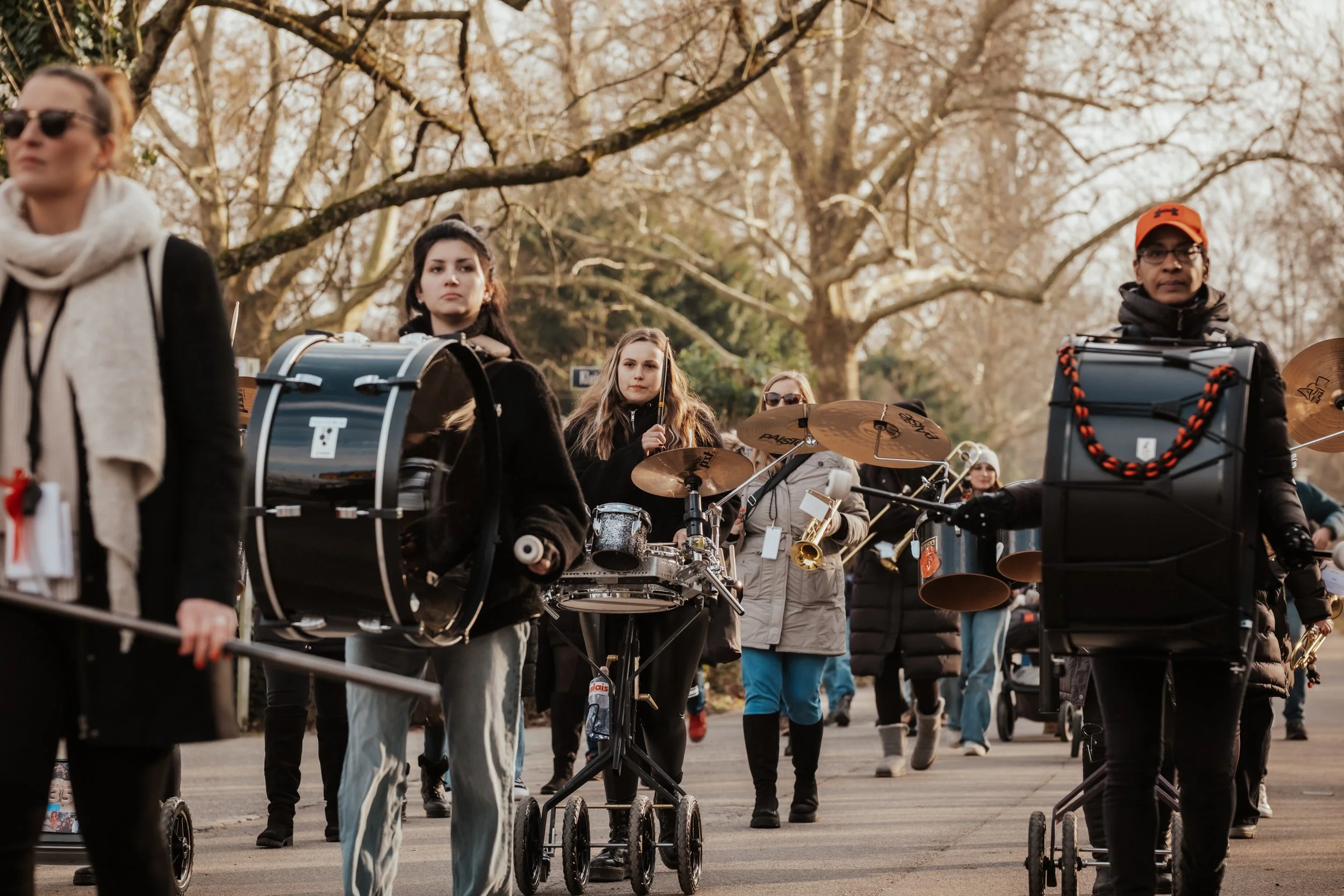 Eine Gruppe von Musikern, die draußen in einem Park auf einem Spazierweg gehen, mit verschiedenen Musikinstrumenten, darunter Trommeln, Becken und Saxophon, bei Sonnenuntergang.