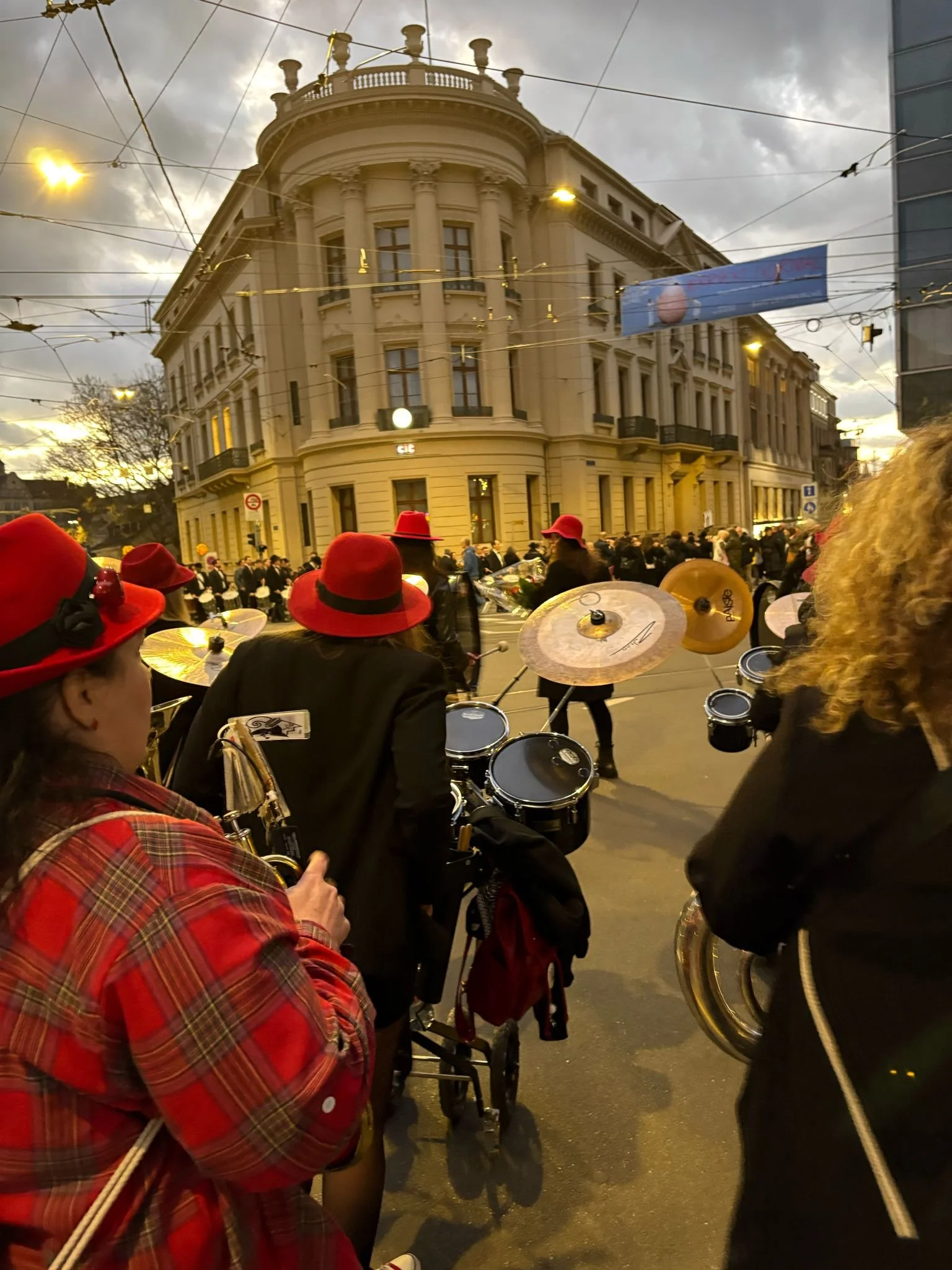 Menschen in roten Hüten und schwarzen Outfits, die auf einer Stadtstraße an einer Parade teilnehmen, mit einem großen, historischen Gebäude im Hintergrund.