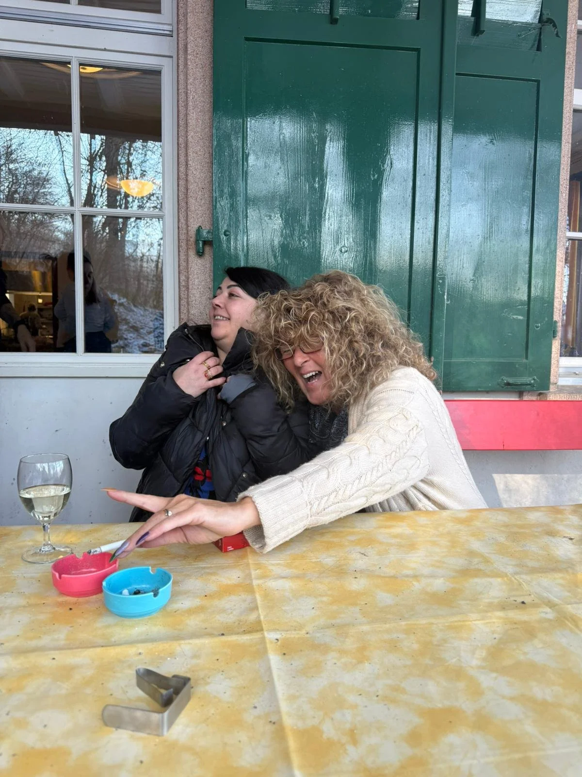 Zwei Frauen lachen und haben Spaß beim Sitzen an einem Tisch im Freien, mit einer Tasse Weißwein, einem roten und einem blauen Aschenbecher auf dem Tisch.