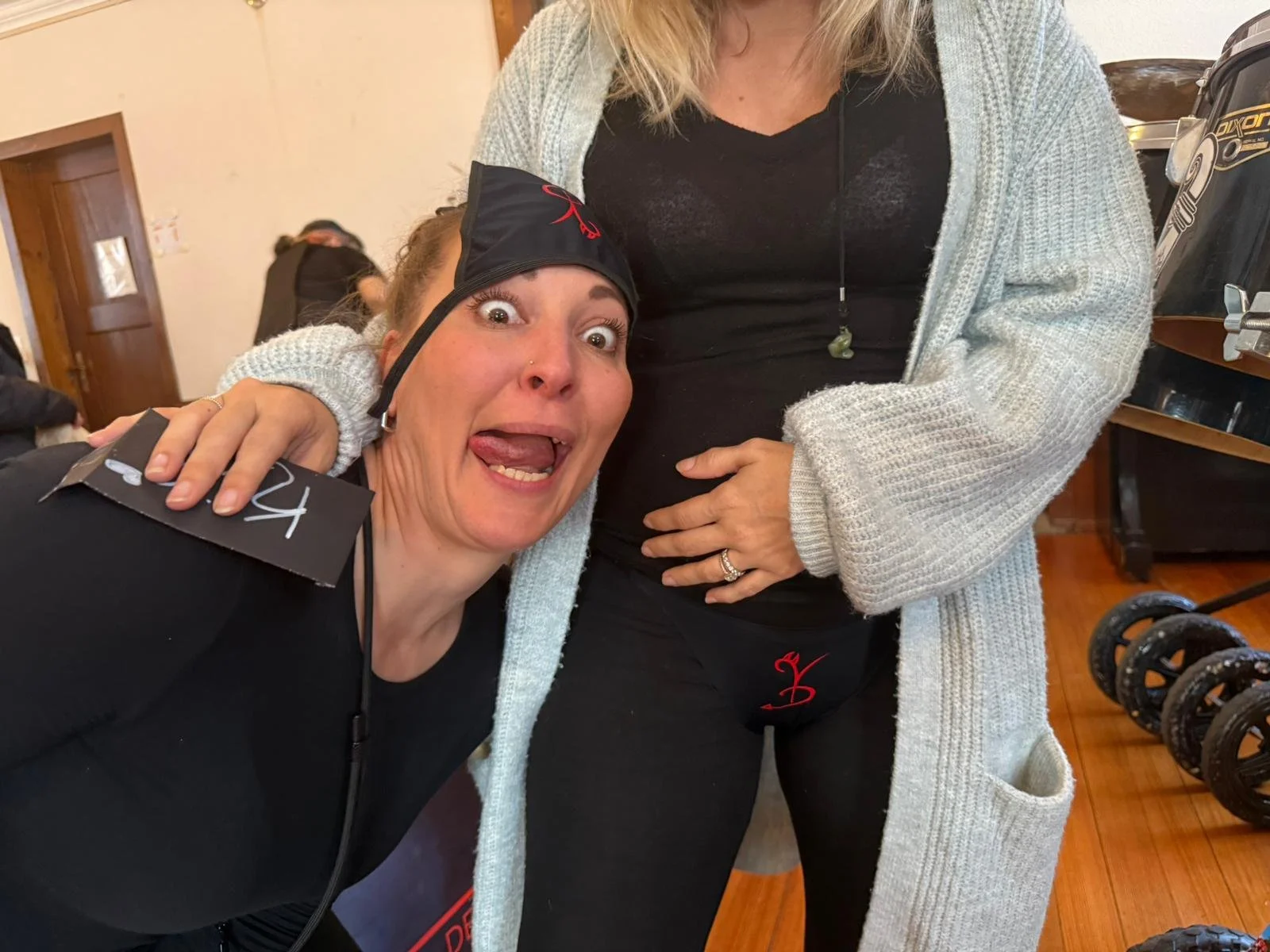 Eine lachende Frau trägt eine schwarze Mütze und ein schwarzes Oberteil, während eine andere Person ihr die Hand auf die Schulter legt. Sie befinden sich in einem Raum mit Holzfußboden und Fitnessgeräten im Hintergrund.