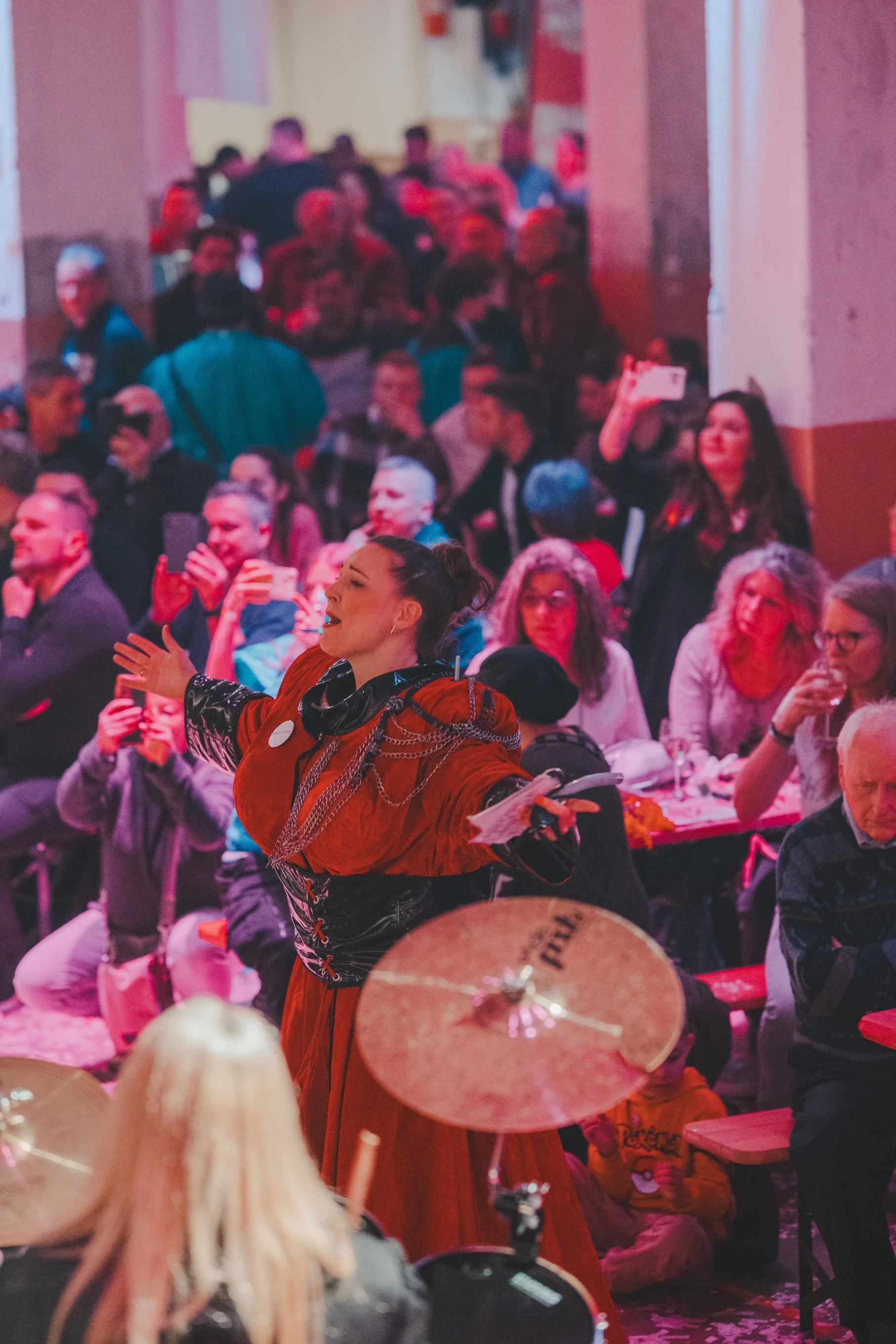 Eine Frau in rotem Kleid mit schwarzen Akzenten und Silberketten auf der Bühne, umgeben von einem Publikum, das sich an Tischen versammelt hat.