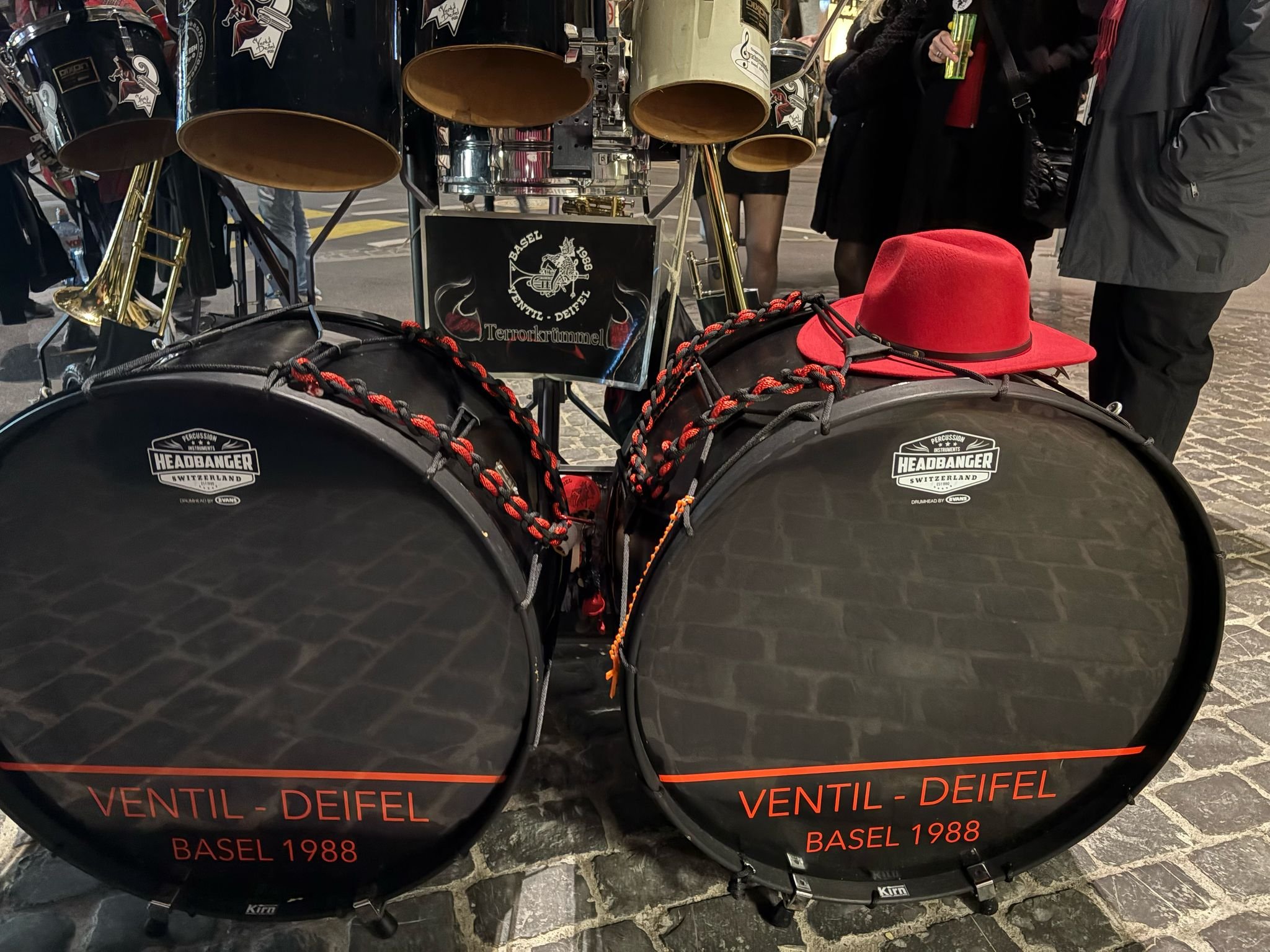 Trommel mit Percussion-Instrumenten, rotkegelige Hut auf den Trommeln, Menschen im Hintergrund, schwizerische Flagge, Text "VENTIL - DEIFEL BASEL 1988" auf den Trommeln, Stadtpflaster, Nachtaufnahme