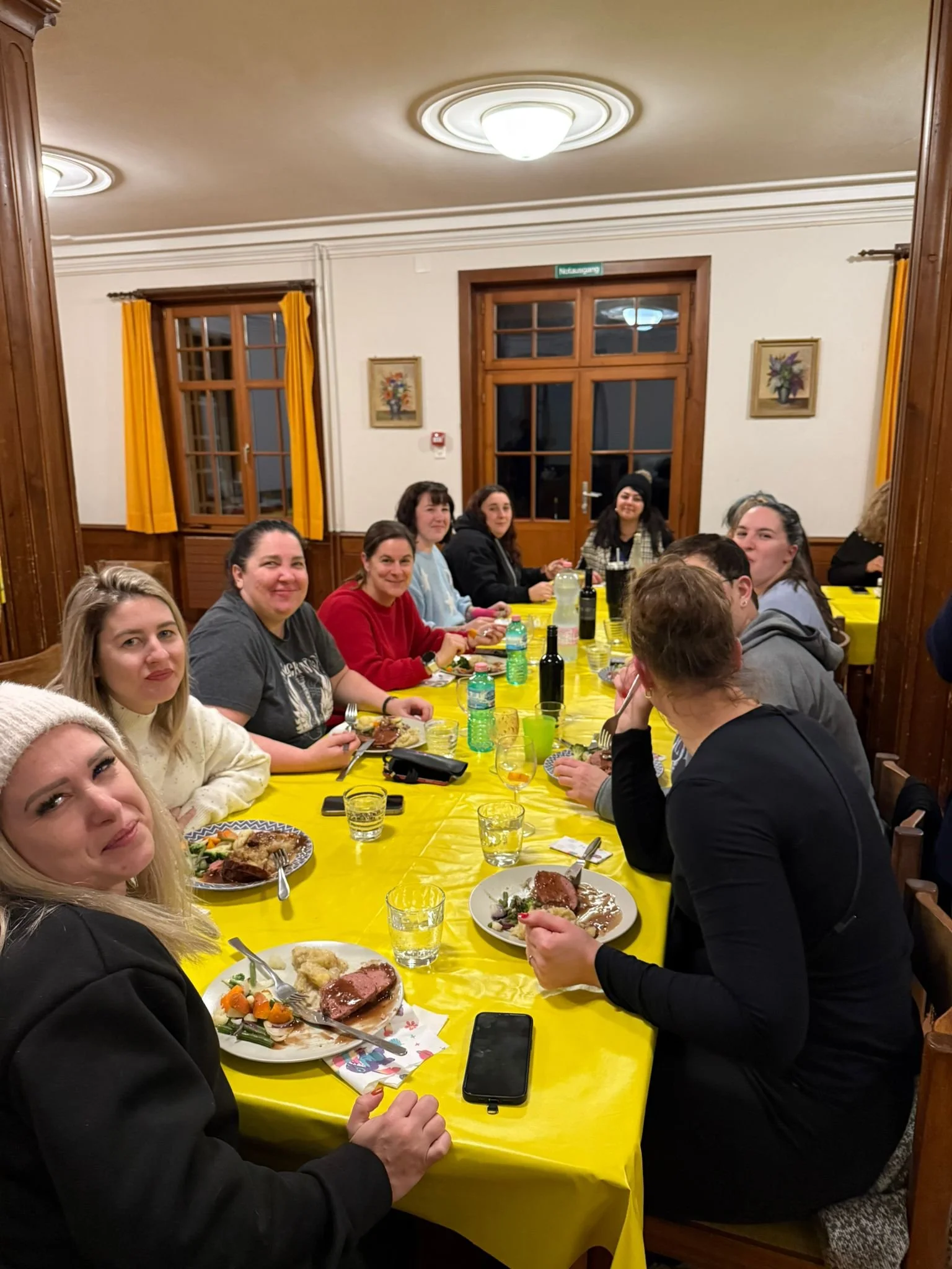 Eine Gruppe von Menschen sitzt an einem langen Tisch mit gelber Tischdecke bei einem gemeinsamen Essen in einem rustikalen Raum mit Holzfenstern und gelben Vorhängen.