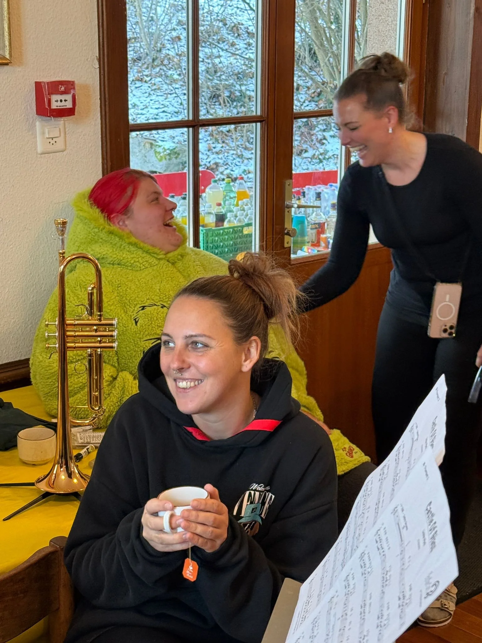 Drei Frauen lachen und unterhalten sich in einer gemütlichen Küche, eine sitzt mit einer Tasse in der Hand, eine andere steht und greift sie am Arm, während im Hintergrund eine weitere Person unter einer Decke sitzt.