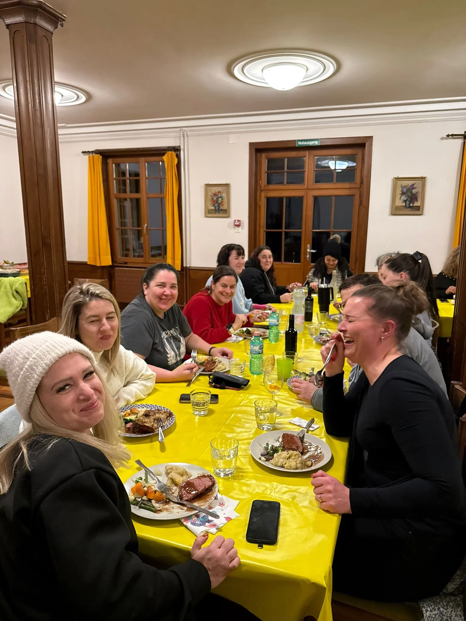 Menschen sitzen an einem langen Esstisch mit gelber Tischdecke und essen gemeinsam in einem Raum mit Holzfenstern und gelben Vorhängen, lachen und unterhalten sich.