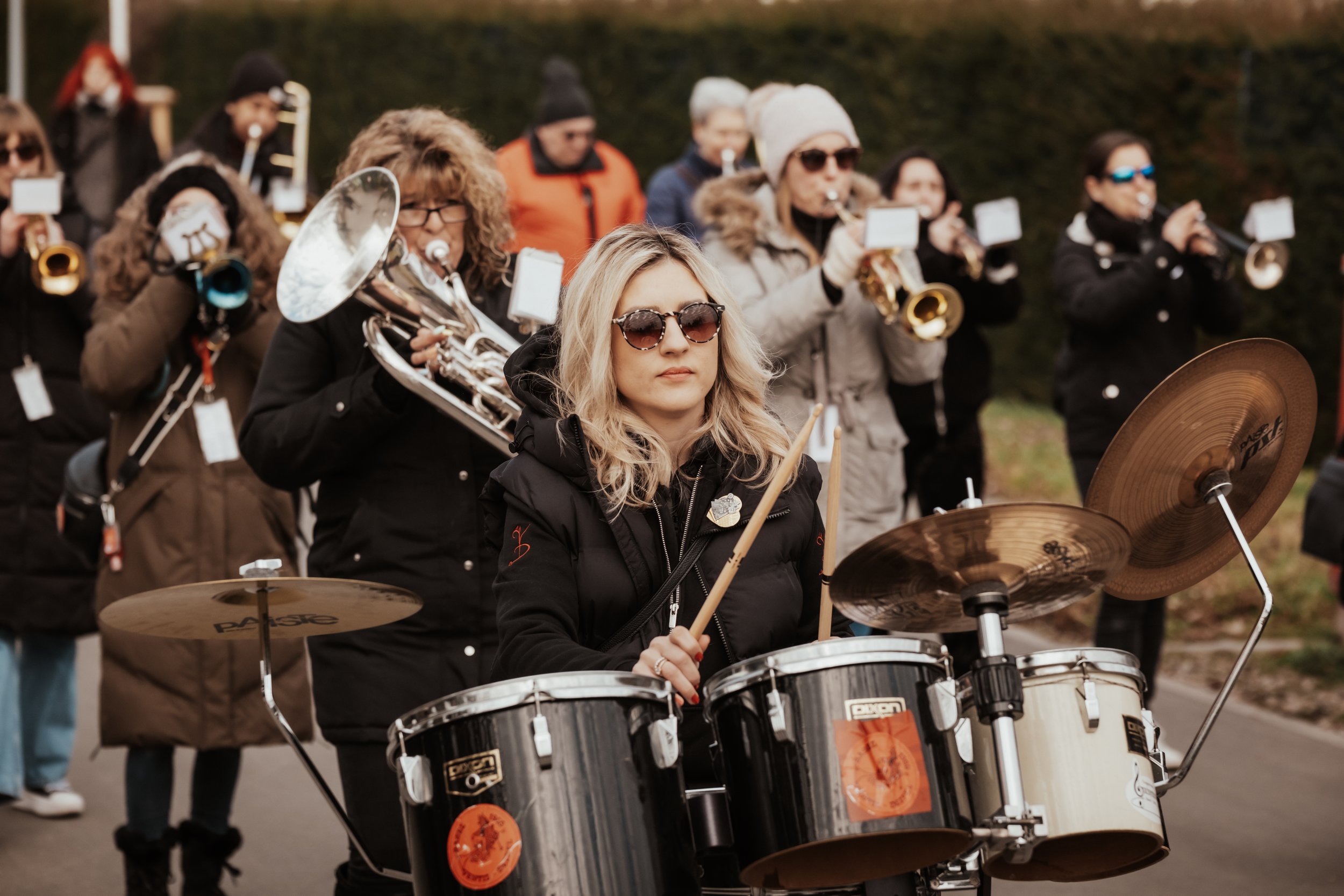 Eine junge Frau spielt Schlagzeug, während eine Gruppe von Musikern im Hintergrund Blasinstrumente spielt, alle tragen Winterkleidung bei einem Musikfest im Freien.