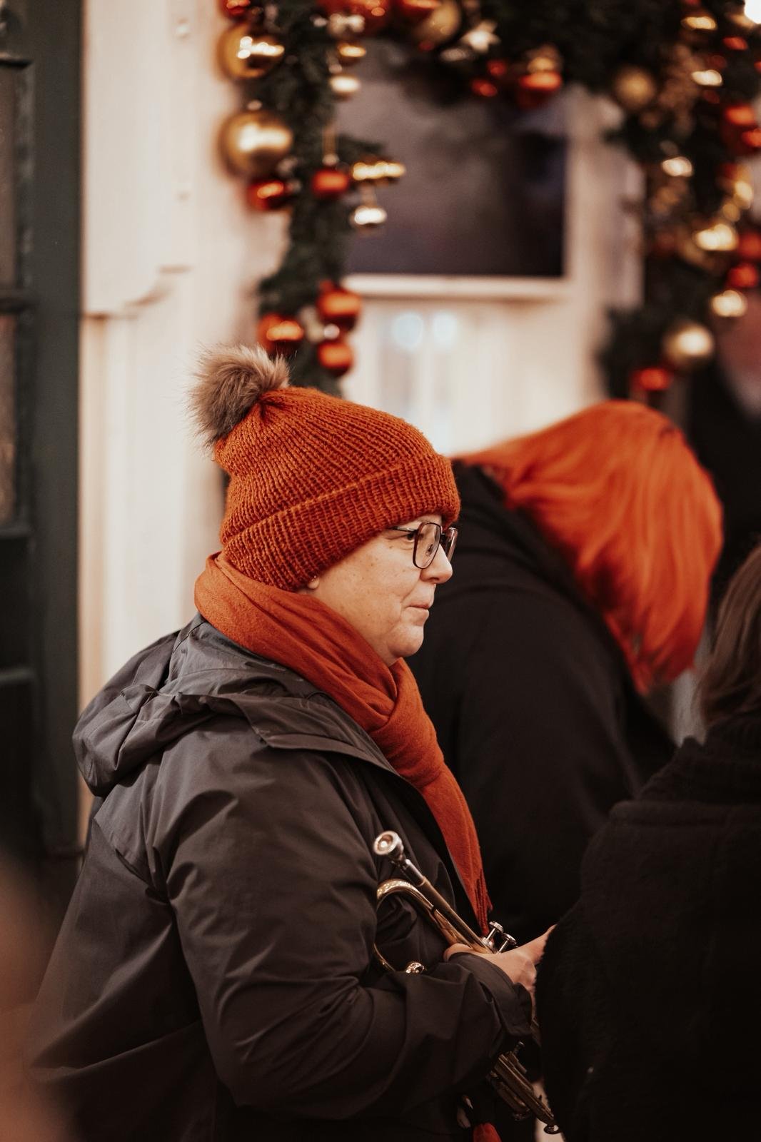 Frau mit rotem Winterhut und Brille hält eine Flöte in der Hand bei einer festlichen Gelegenheit im Inneren, mit Weihnachtsdekoration im Hintergrund.