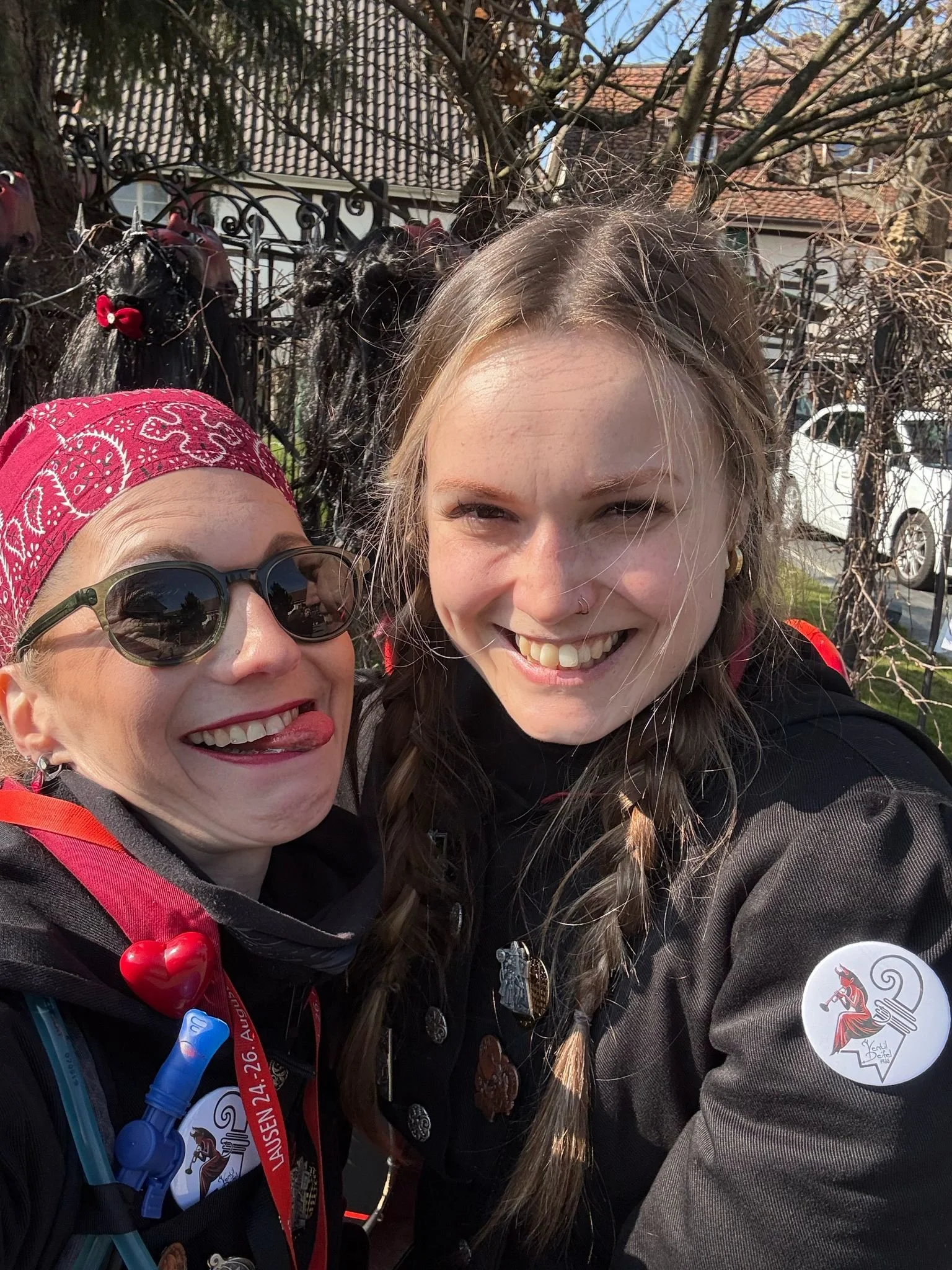 Zwei lachende Frauen im Freien, eine trägt eine rote Kopftuch mit weißem Muster, Sonnenbrille, der anderen sind zwei geflochtene Haare, sie trägt ein schwarzes Jacke mit Aufklebern, im Hintergrund Bäume und Häuser.