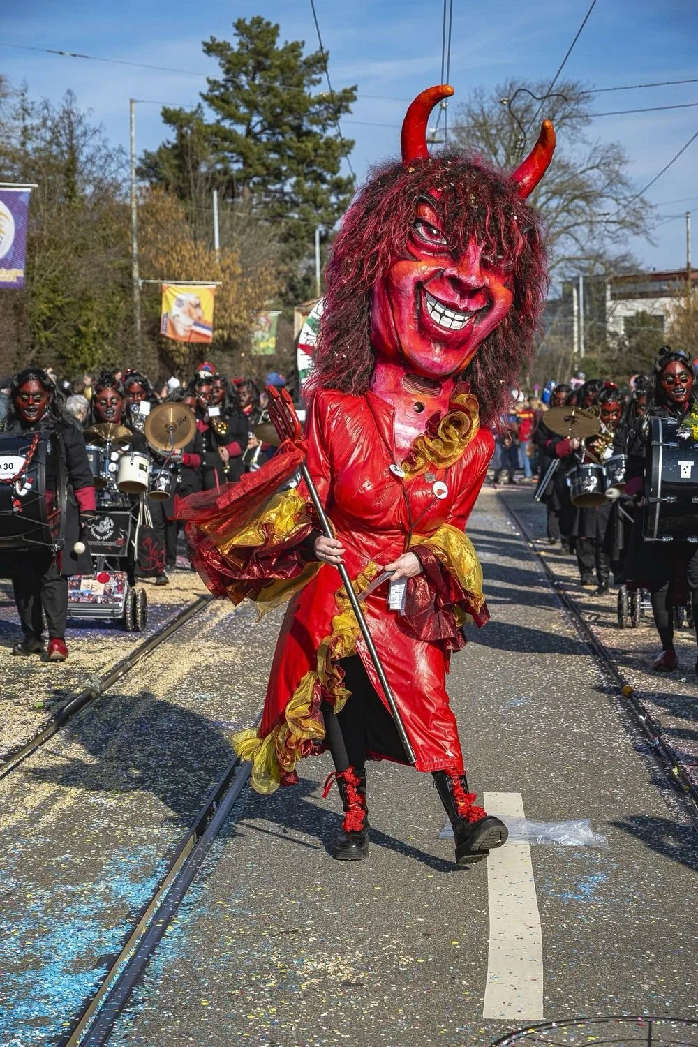 Person in aufwendigem rot-schwarzen Teufelskostüm bei Karnevalsparade, umgeben von Musikkapelle in ähnlichen Kostümen, bunte Konfetti auf der Straße, sommerlicher Himmel.