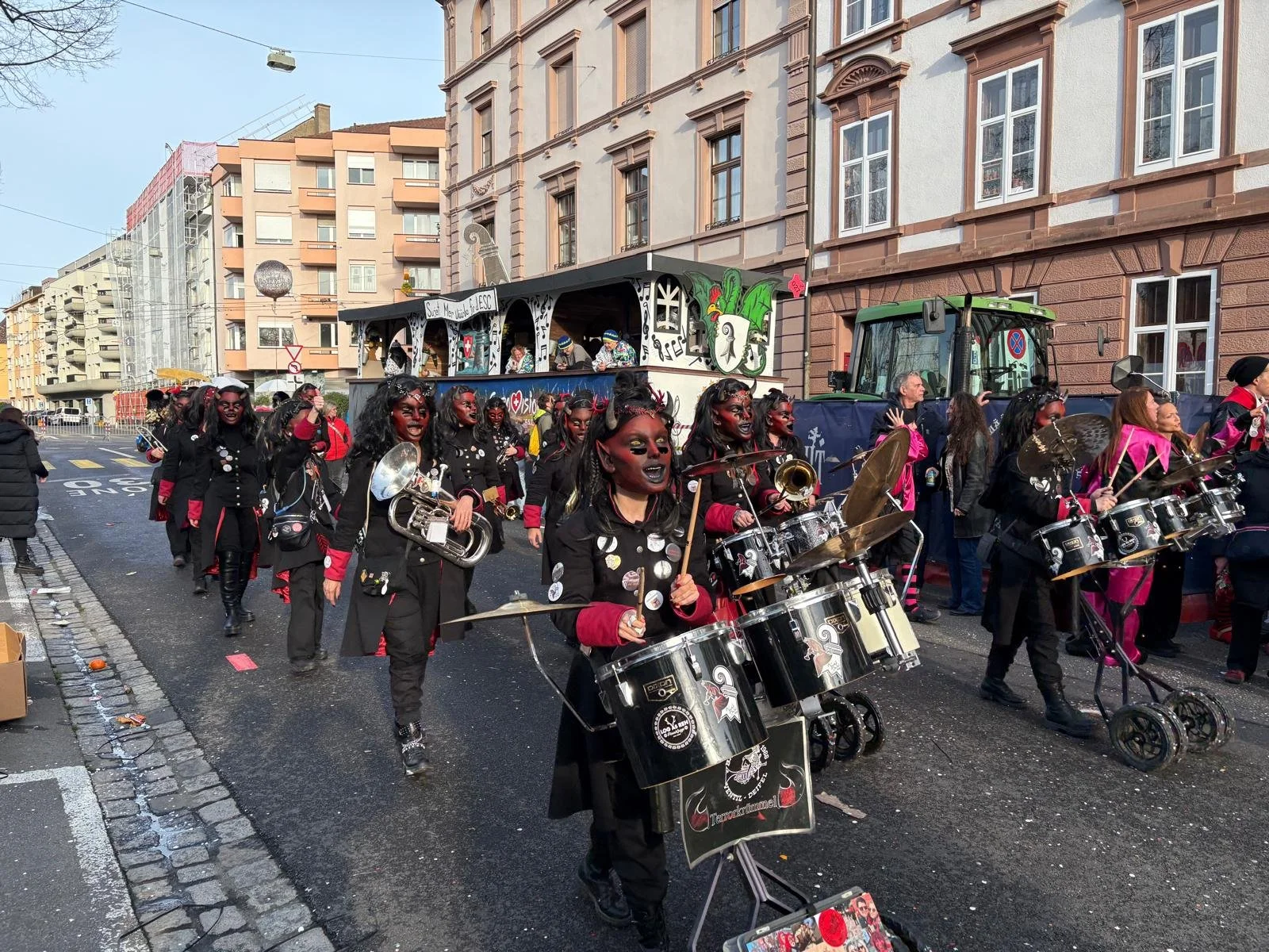 Eine Gruppe von Menschen in schwarzen Kostümen mit roten Akzenten bei einem Umzug, die Musik spielen, während sie die Straße entlang ziehen, auf einer Stadtstraße mit mehrstöckigen Gebäuden im Hintergrund.