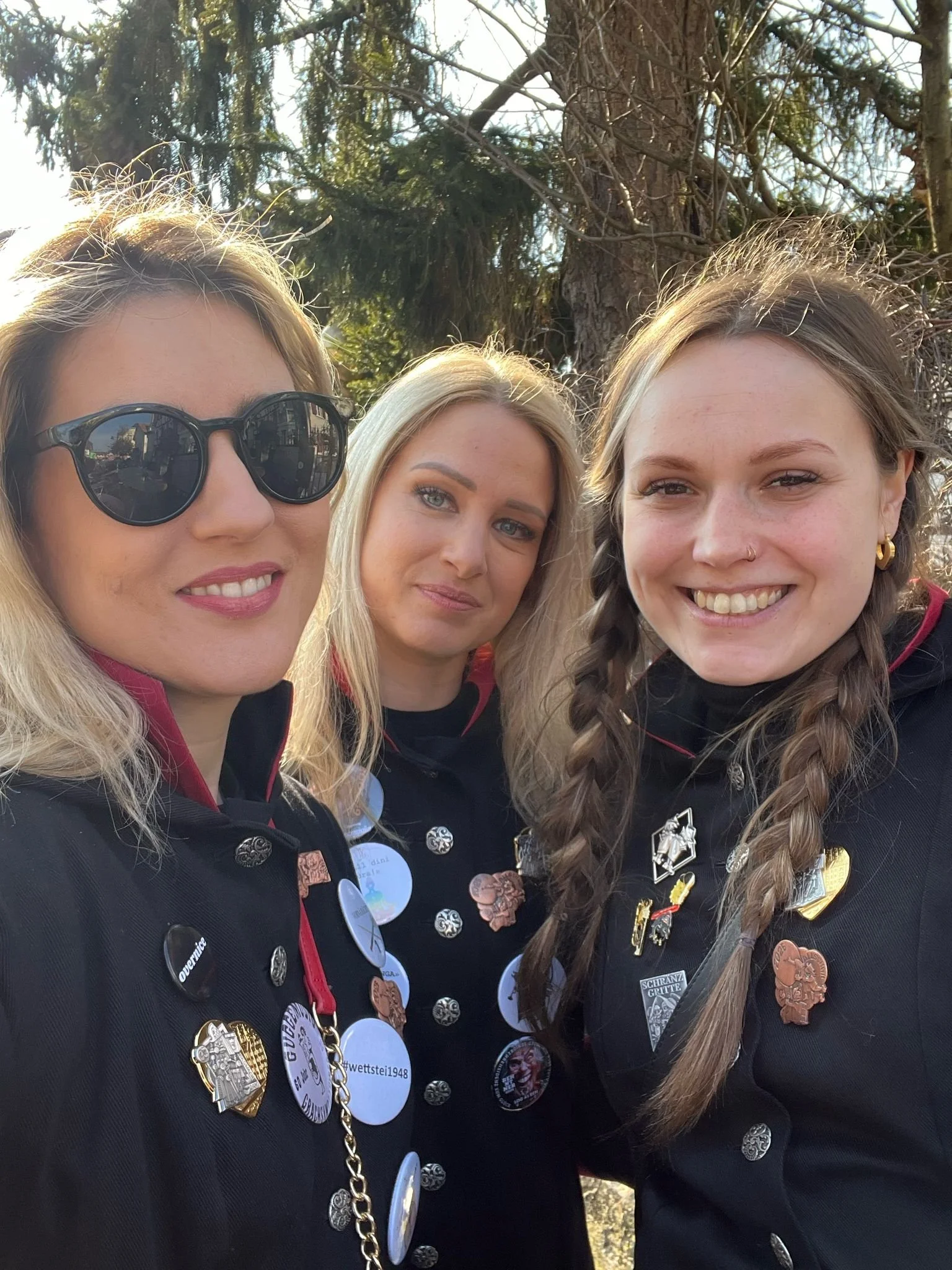 Drei Frauen in schwarzer Uniform mit Anstecknadeln und Buttons, stehen vor einem Baum im Freien, Sonnenlicht, lachend, zusammen für ein Gruppenfoto.