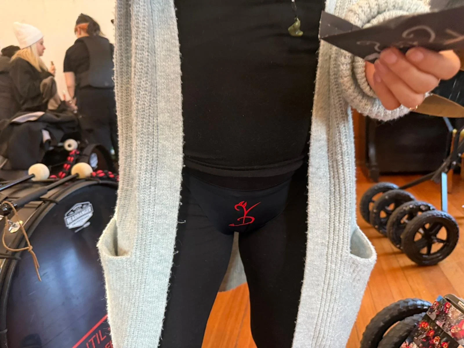 Nahaufnahme einer Person mit grauem Mantel, schwarzer Kleidung und einer schwarzen Unterhose mit rotem Logo, die in einem Raum mit anderen Menschen und Sportgeräten steht.