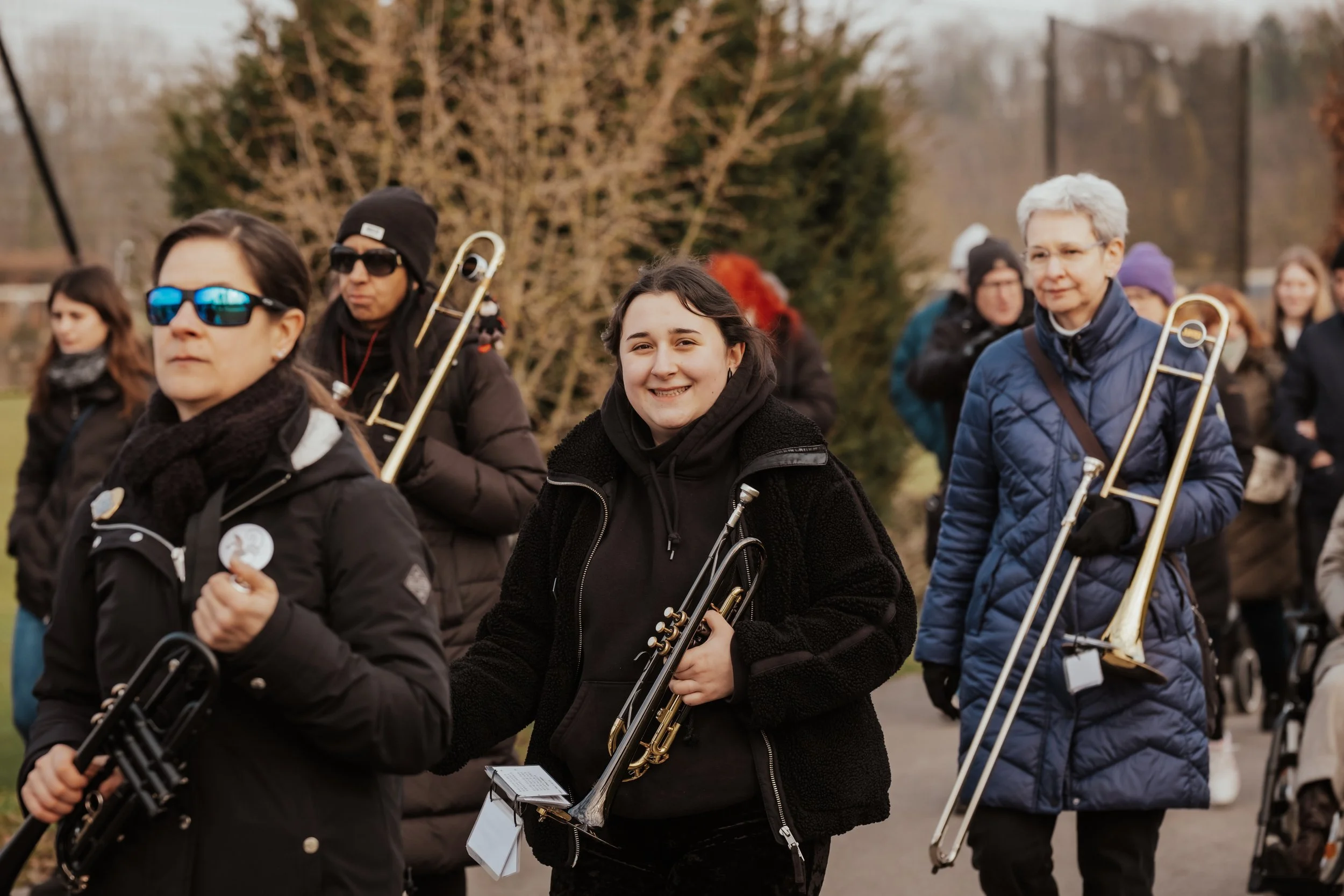 Gruppe von Menschen beim Musizieren im Freien, einige halten Trompeten und andere Posaunen, im Hintergrund Bäume und eine unklare Gruppe von Personen.