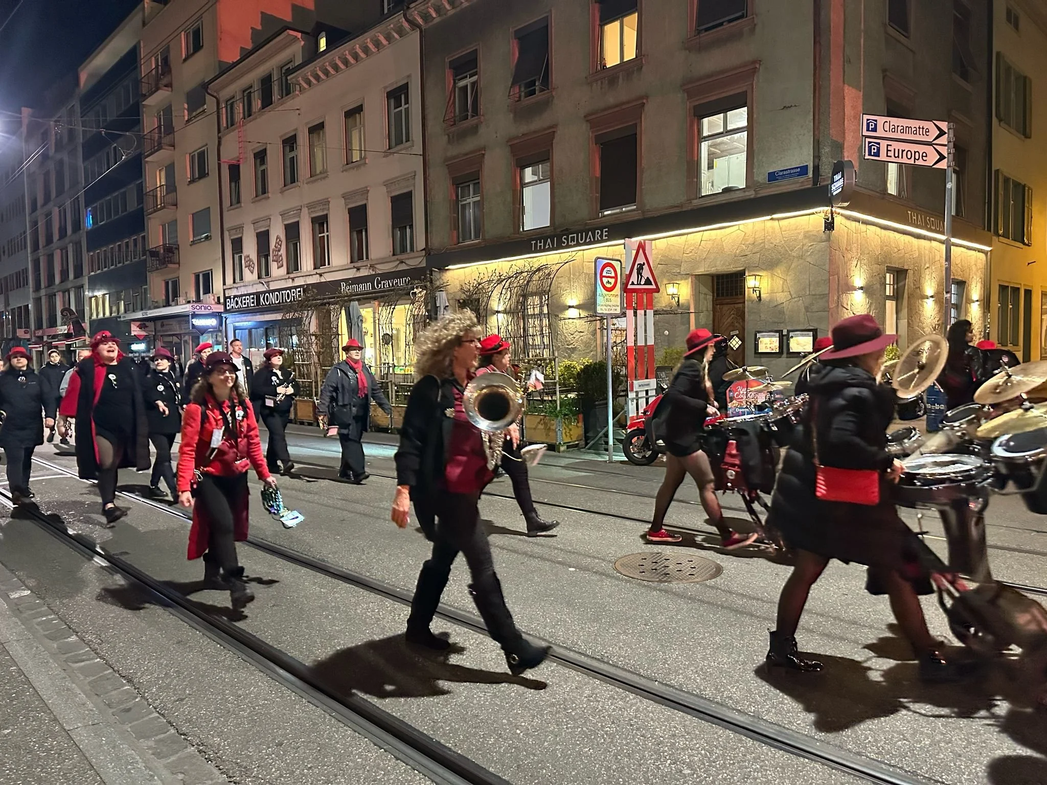 Eine Gruppe von Menschen, die in einer Stadtstraße abends auf einer Tramspur marschieren, einige in uniformähnlichen schwarzen Outfits mit roten Akzenten und Hüten, einige mit Musikinstrumenten, wie Trommeln und Saxophonen.