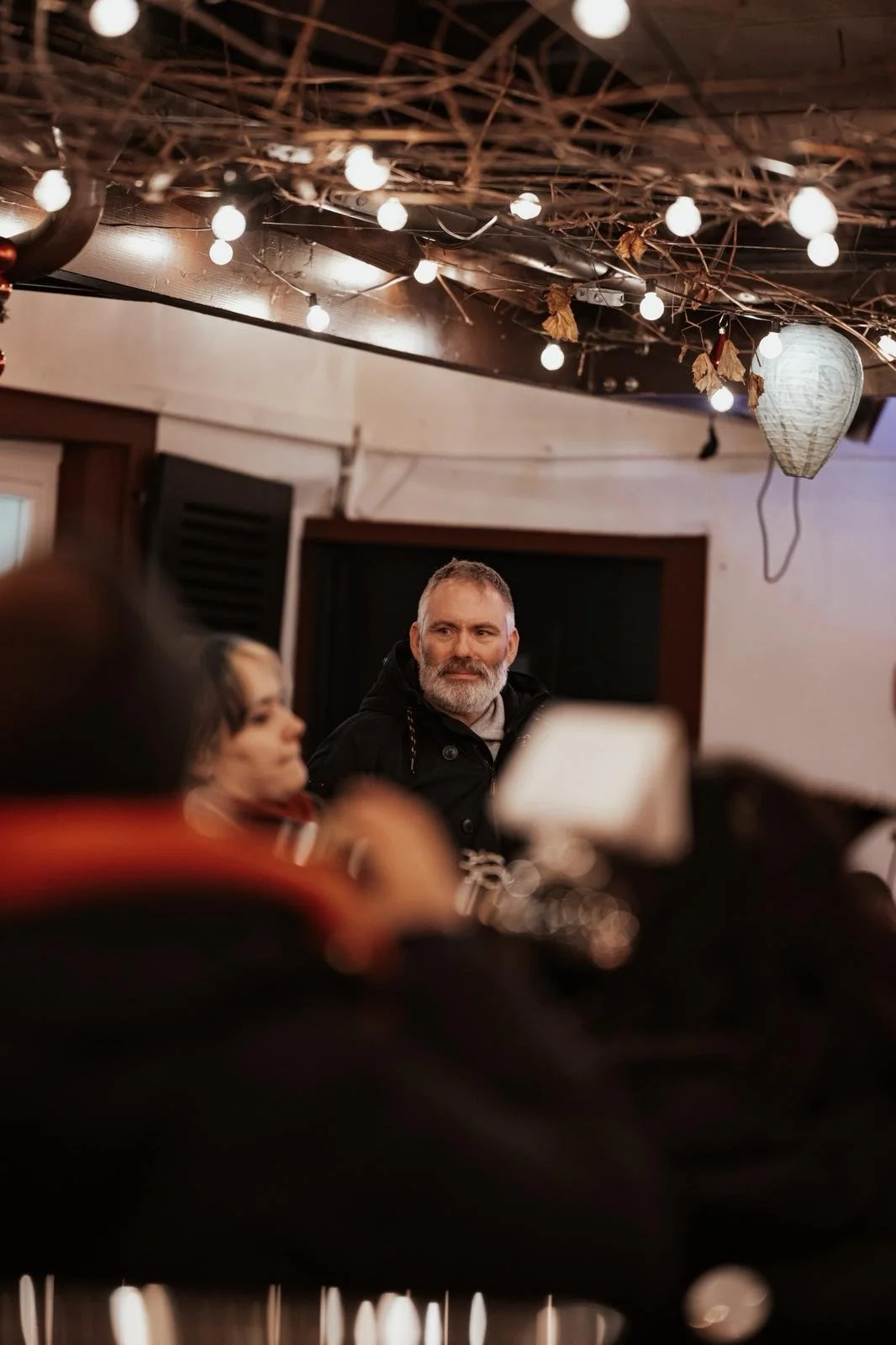 Ein Mann mit grauem Bart sitzt in einem gemütlichen Raum, umgeben von Menschen, bei einer Veranstaltung oder einem Treffen, mit warmem Lampenlicht und Dekoration an der Decke.