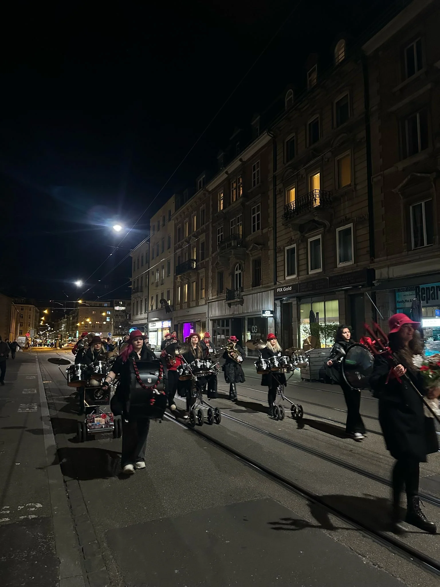 Eine Gruppe von Menschen, die in der Nacht eine Straßenbahnlinie entlang marschieren, alle tragen schwarz und rote Kleidung und Hüte, einige sind mit Musikinstrumenten und Paketen ausgestattet.