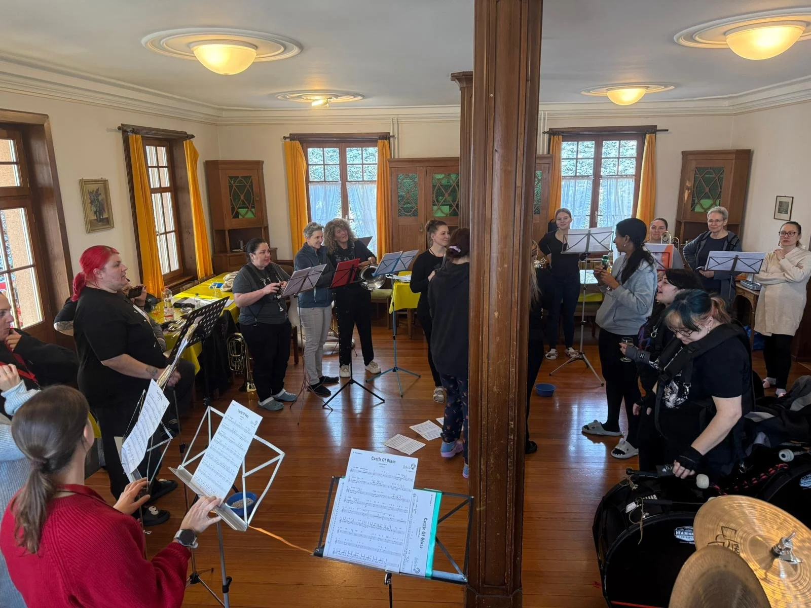 Musikerinnen und Musiker proben in einem Raum. Manche spielen Blasinstrumente, andere halten Notenblätter in den Händen.