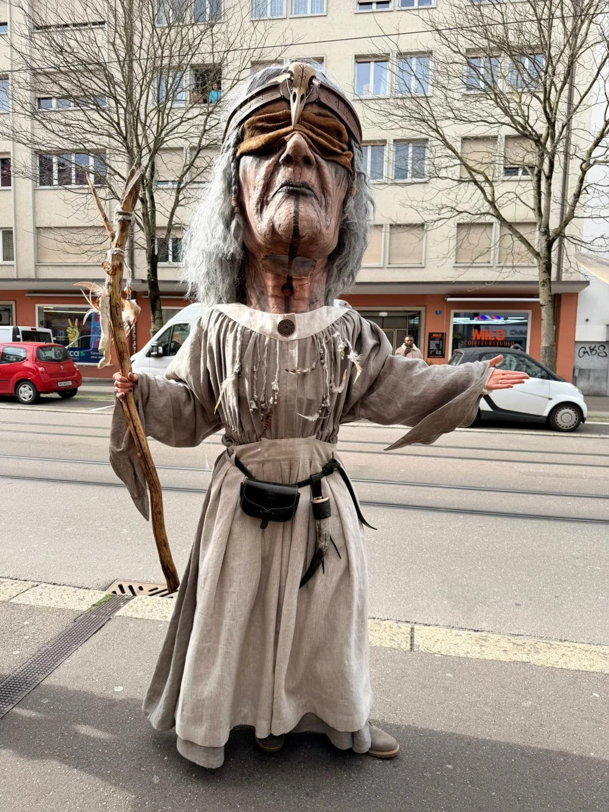Person in einem Kostüm, das eine alte, weise Figur darstellt, trägt ein langes graues Gewand, eine Augenbinde und hält einen Zauberstab, steht auf einer Straße in einer Stadt.