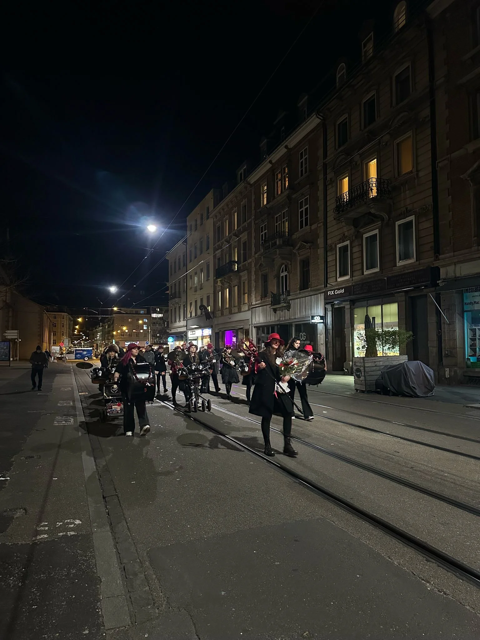 Menschen mit Blumen und Musikinstrumenten, die nachts auf einer Straße mit Bahngleisen gehen, in einer Stadt mit mehrstöckigen Gebäuden.