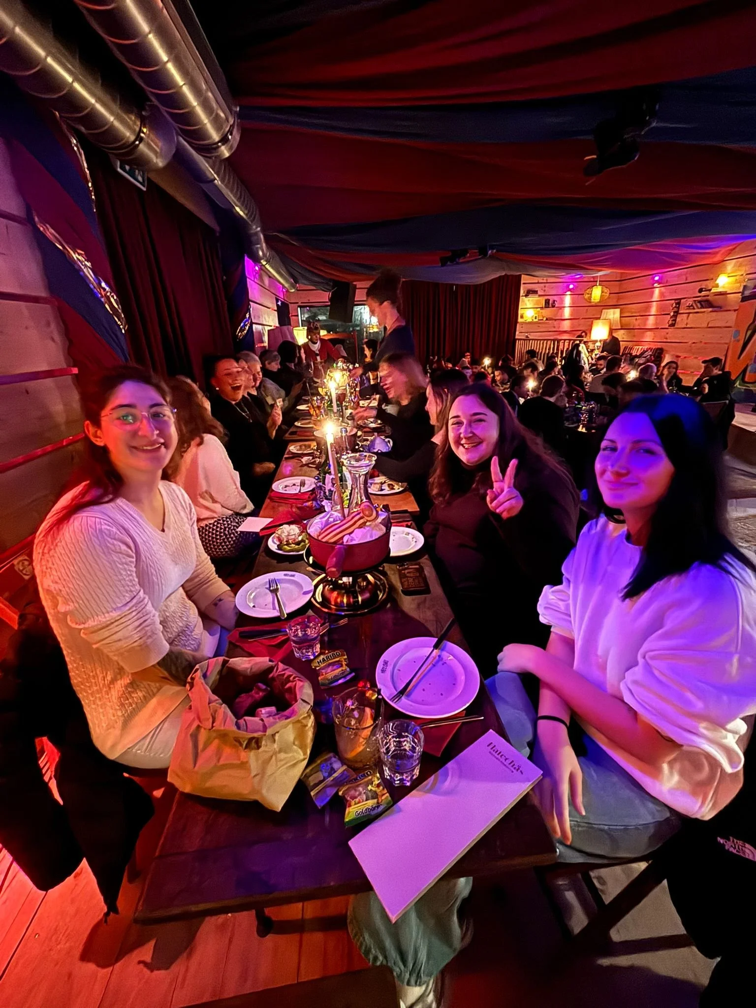 Menschen sitzen bei einem Abendessen in einem dekorierten, gemütlichen Raum mit buntem Beleuchtungskonzept und lachen.