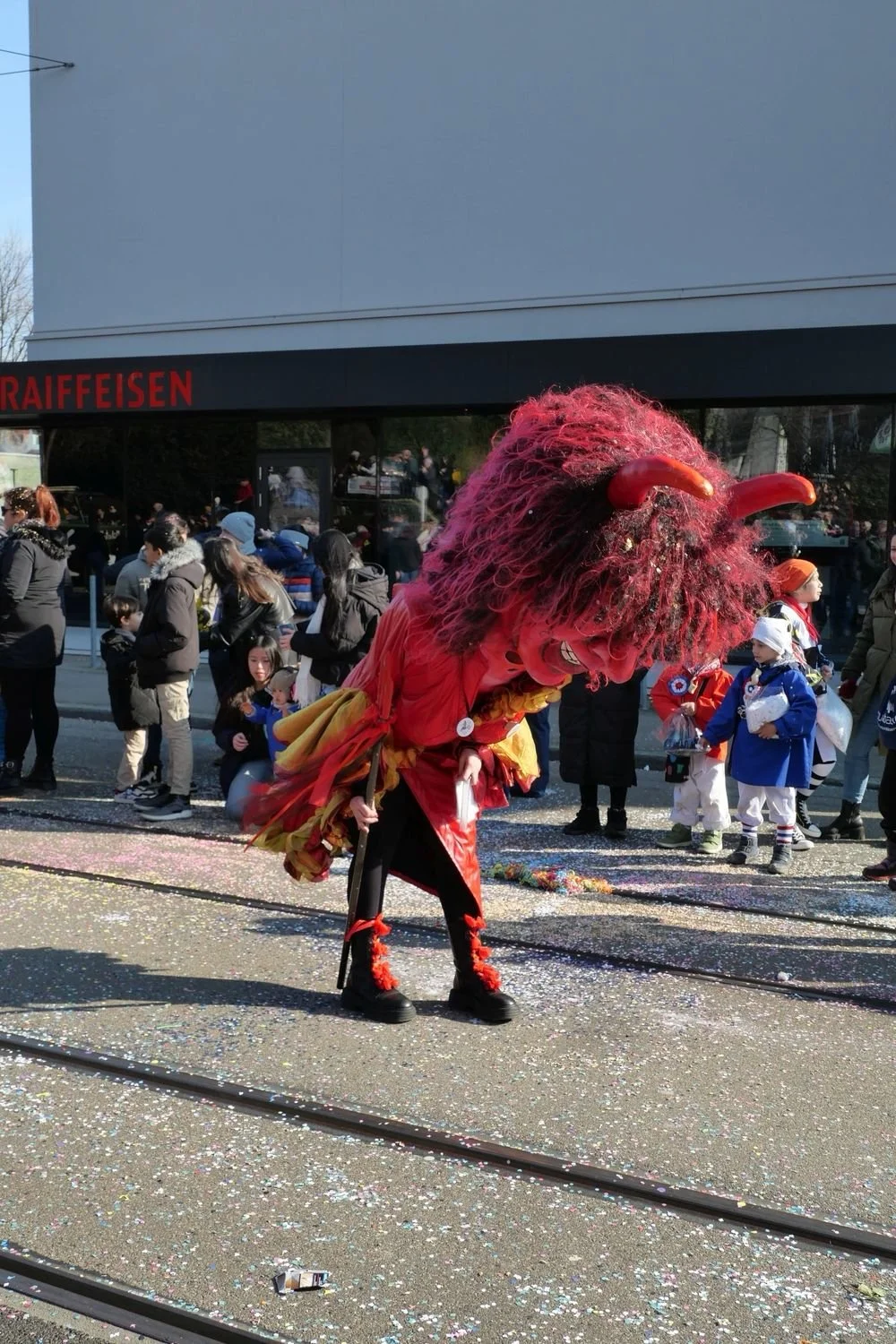Ein fantastisches Maskottchen in einem Karneval oder Fasnachtsumzug, mit einem großen roten und rosa Kopf, großen Hufen, und einer bunten Kleidung, umgeben von einer Menge Menschen und Kindern, die an einem sonnigen Tag feiern.
