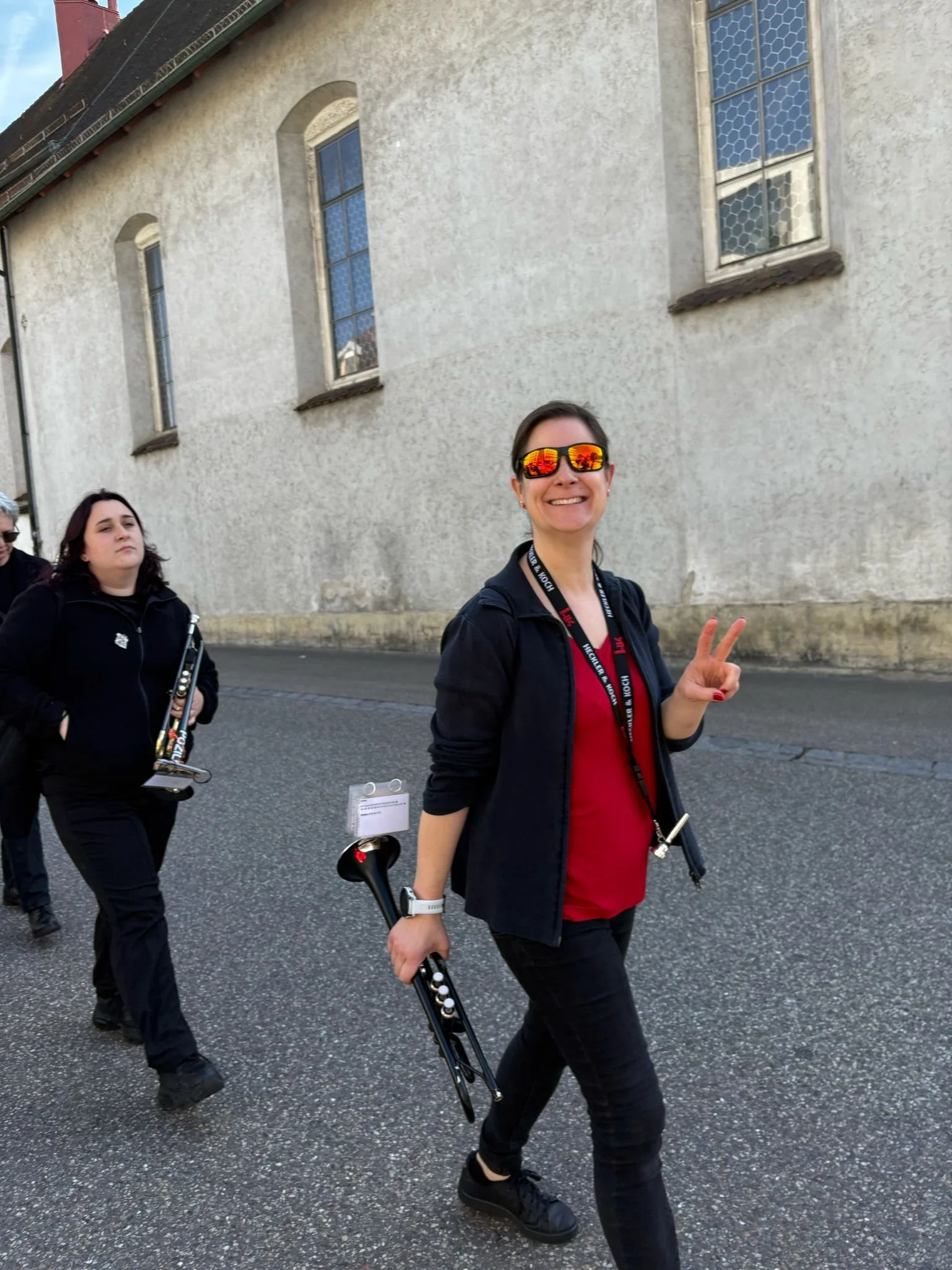 Junge Frau lächelt, hebt friedlich ihre Hand in Peace-Zeichen, trägt reflektierende Sonnenbrille, schwarze Kleidung und hält ein Schloss in der Hand, während sie mit anderen Menschen an einem Tag spazieren geht.
