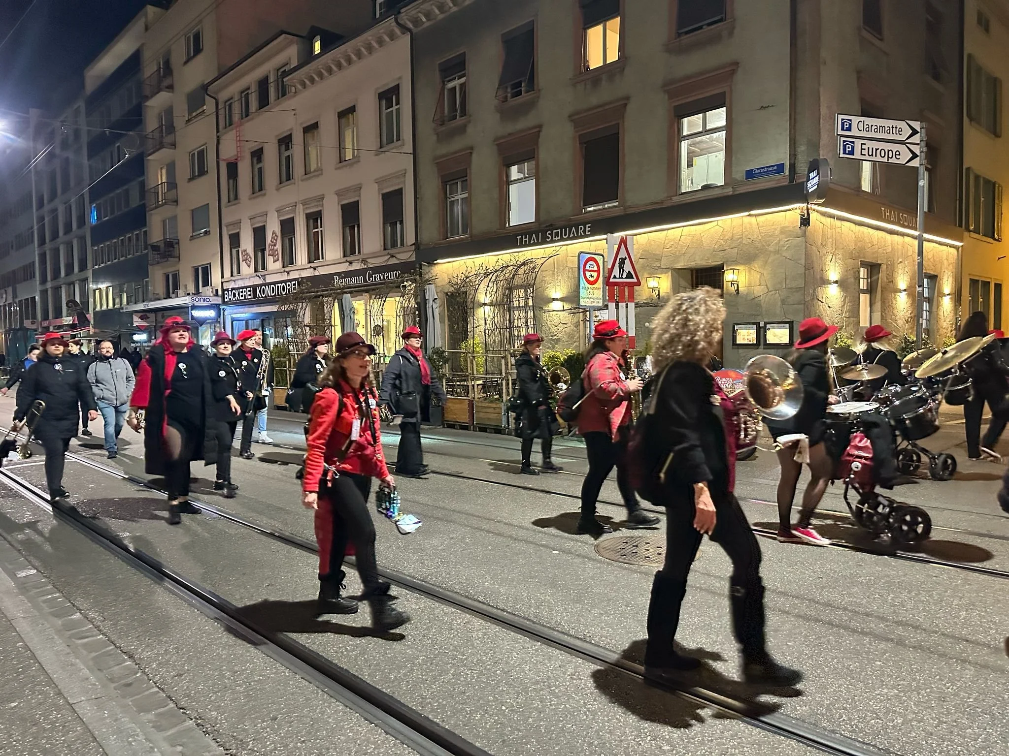 Menschen ziehen nachts in einer Stadtstraße mit Straßenbahnschienen, sie tragen rote Hüte und schwarze Kleidung, einige spielen Musikinstrumente, im Hintergrund sind Gebäude mit beleuchteten Fenstern und Schildern.