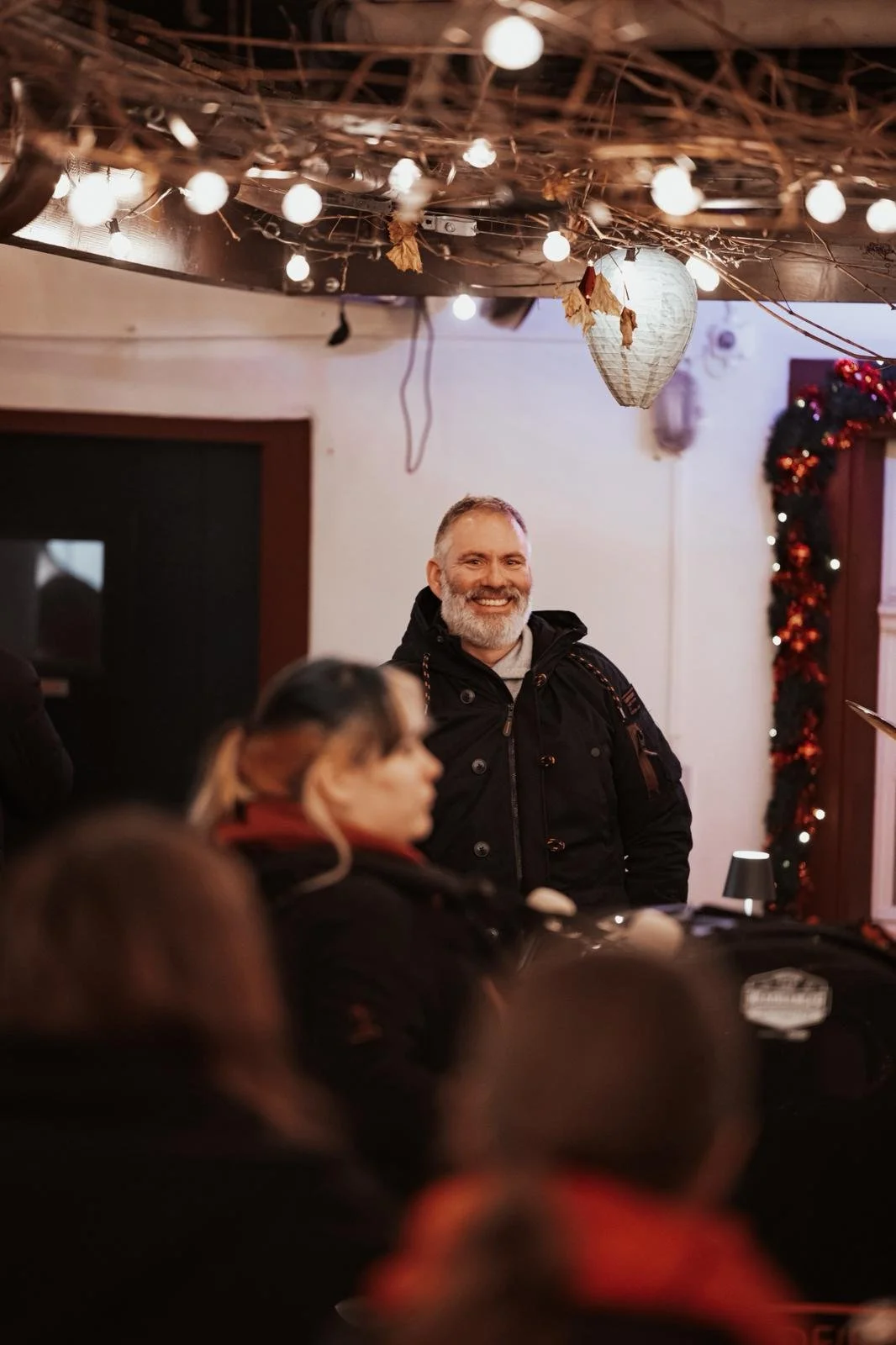 Ein lächelnder Mann mit grauem Bart in einer festlich dekorierten Umgebung, umgeben von mehreren Personen, mit weihnachtlicher Beleuchtung und Dekorationen.