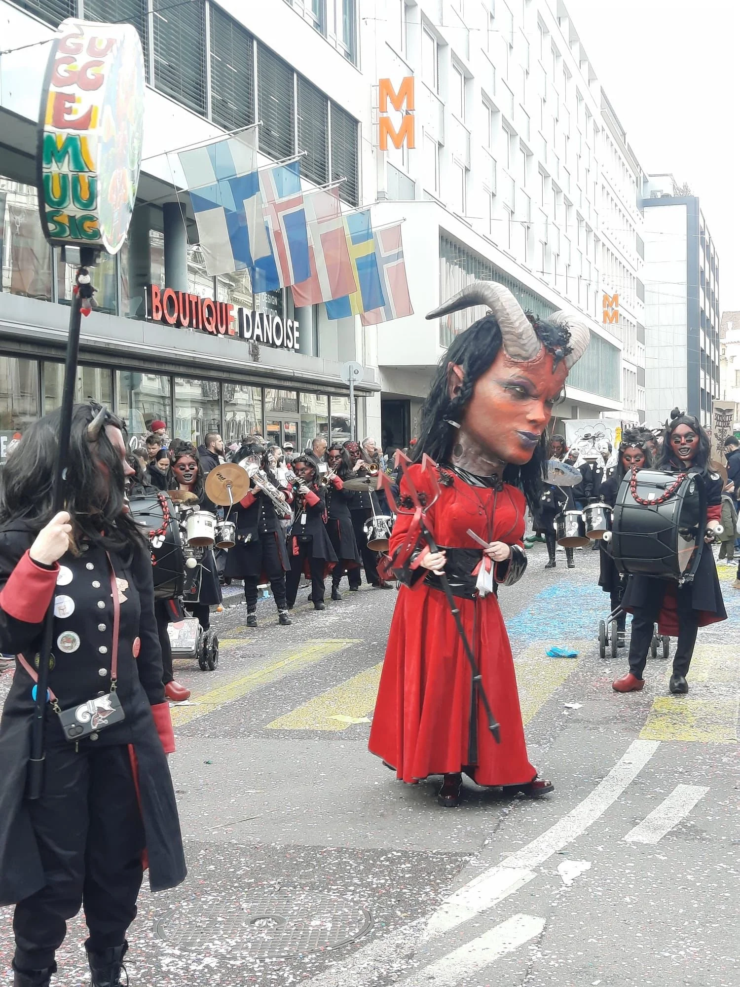 Menschen in Kostümen bei einer Parade, mit einer Person in einem Dämonenkostüm mit großen Hörnern und roten Kleid im Mittelpunkt.