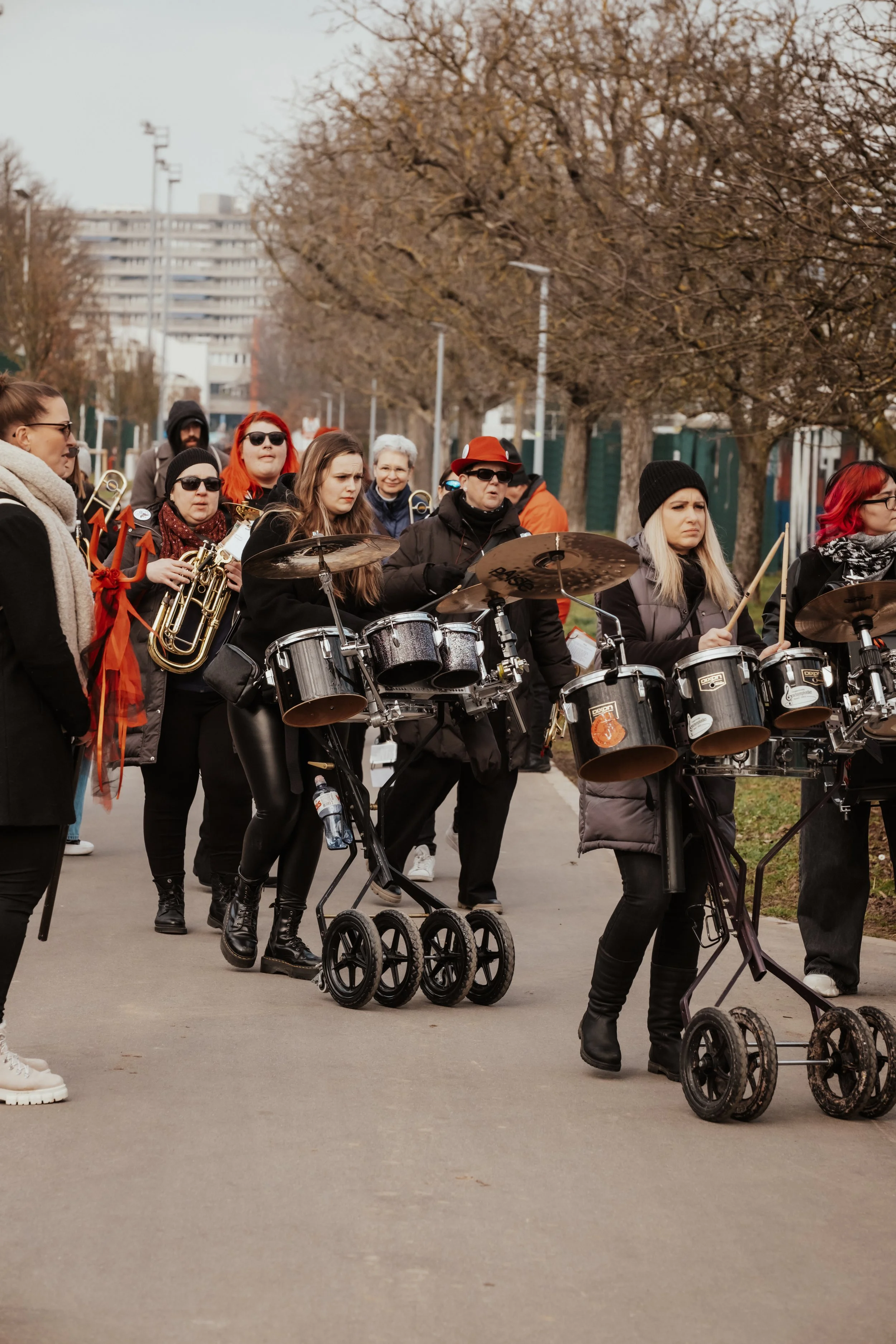Gruppe von Menschen, die eine Straßenmusik- oder Demonstrationsveranstaltung mit Musikinstrumenten wie Trommeln und Blechbläsern durchführen, bei kaltem Wetter auf einer Straße mit Bäumen und Gebäuden im Hintergrund.
