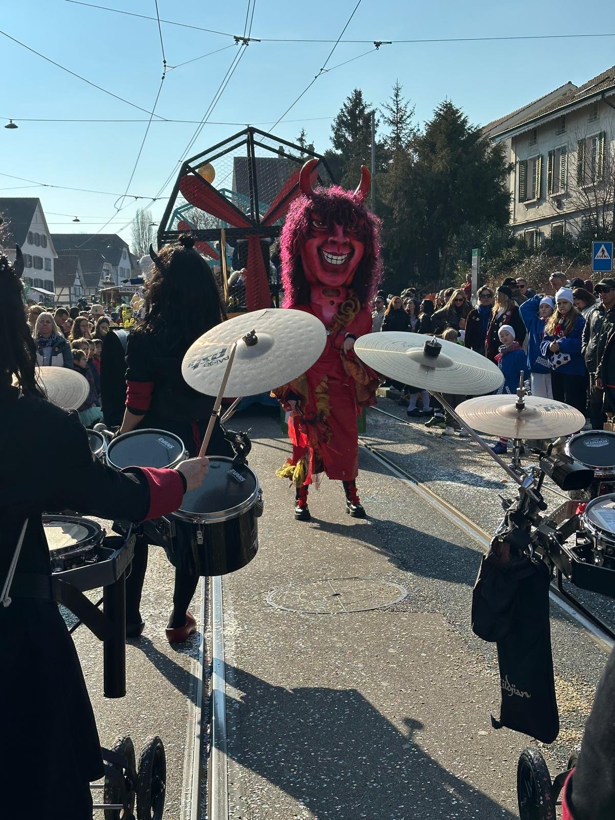 Karnevalsfigur auf einem Festwagen, umgeben von Menschen, auf einer Straße mit Tramgleisen, bei sonnigem Wetter.