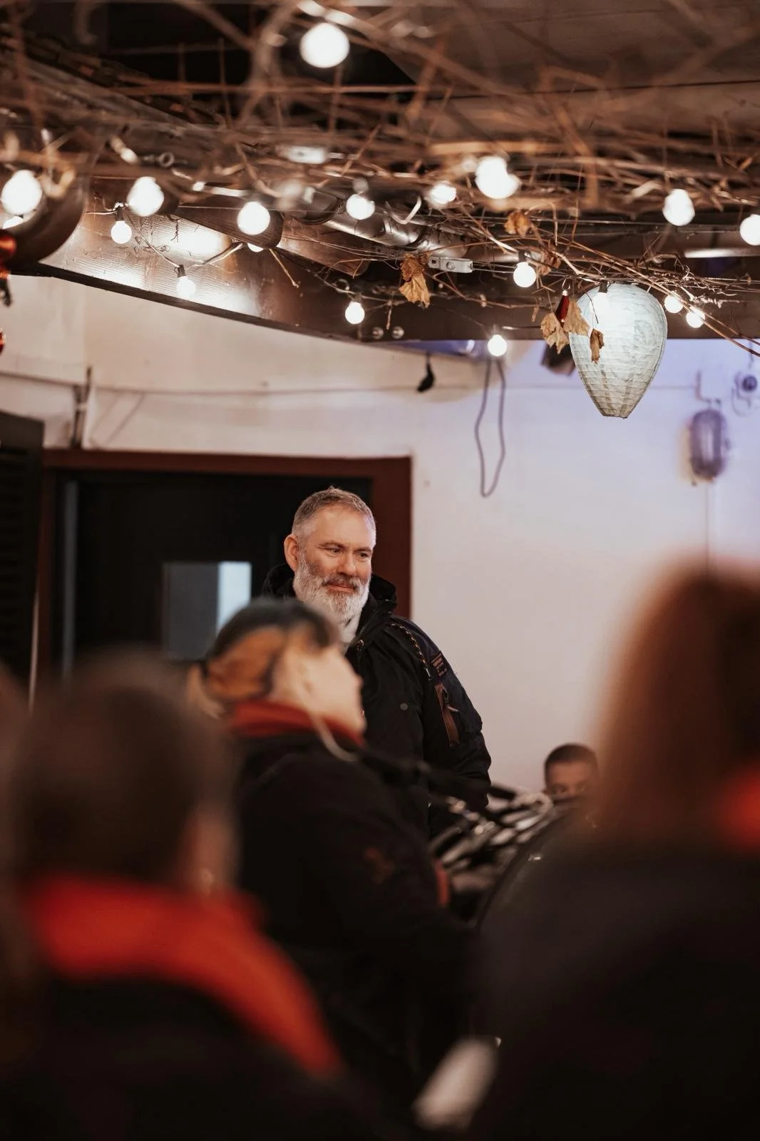 Ein Mann mit grauem Bart steht in einem Raum mit mehreren Menschen, die ihm zuhören. Über ihm hängen Lichterketten und Dekorationen. Es scheint eine gemütliche Atmosphäre zu sein.