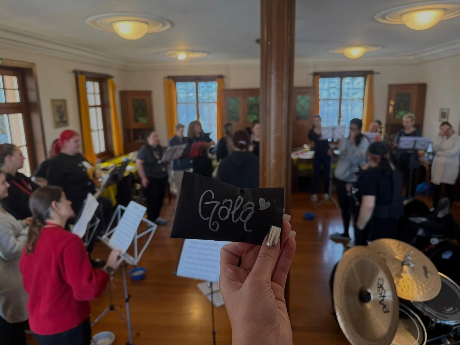 Ein Chor probt in einem Raum mit Holzfußboden, großen Fenstern und gelben Vorhängen. Eine Hand hält ein kleines schwarzes Schild mit dem Wort 'Gala' und einem Herzsymbol, im Vordergrund sind Musikinstrumente zu sehen.