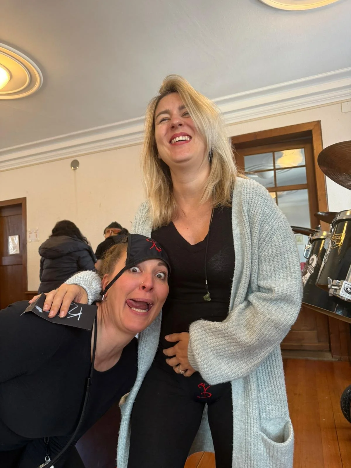 Zwei lachende Frauen, eine mit einem schwarzen Cap, die andere trägt eine graue Strickjacke, posieren fröhlich in einem Raum mit Holzarmaturen und einem großen Fenster im Hintergrund.