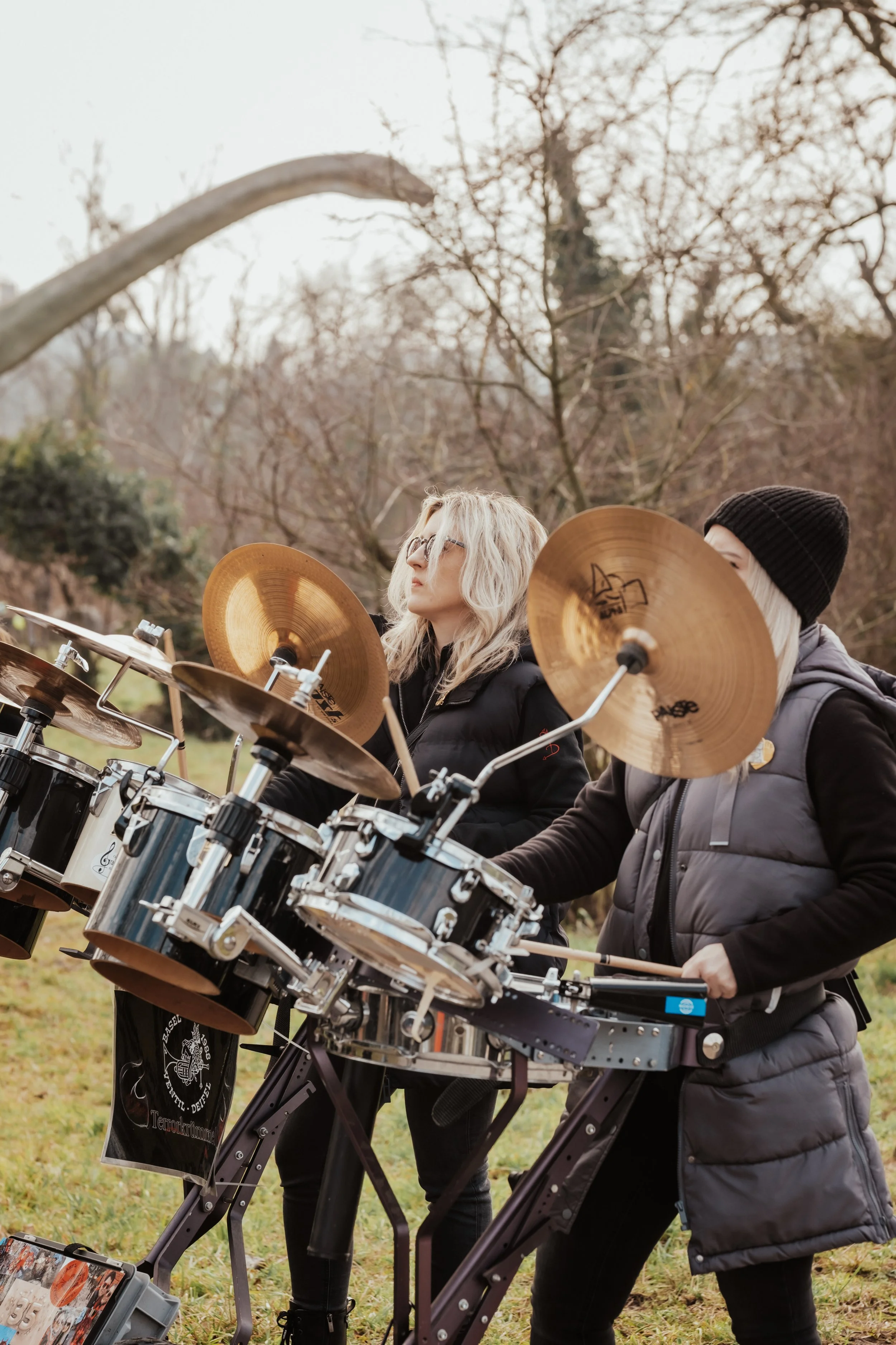 Zwei Menschen spielen auf einem elektronischen Schlagzeug im Freien, umgeben von Bäumen mit kahlen Zweigen, vermutlich im Herbst oder Winter.