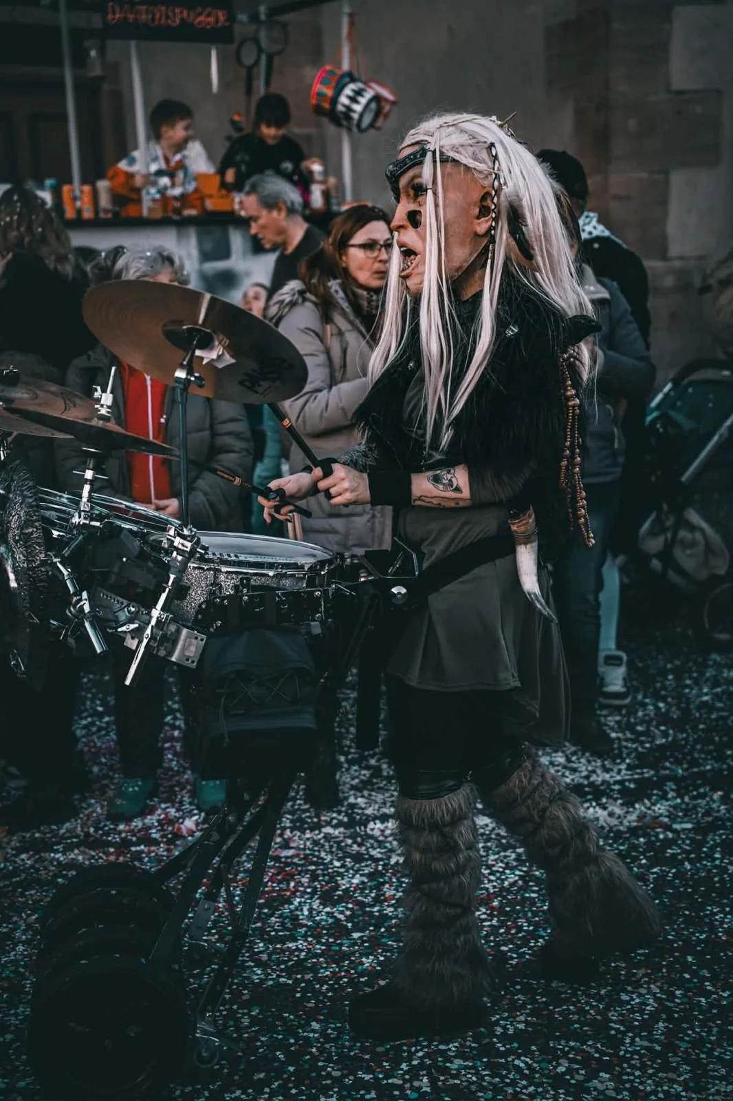 Person in fantasy- oder cosplay-Kostüm mit weißen langem Haar, auf einer Trommel schlagend, bei einer Feier oder Parade, umgeben von mehreren Menschen, im Innenraum, mit Konfetti auf dem Boden.