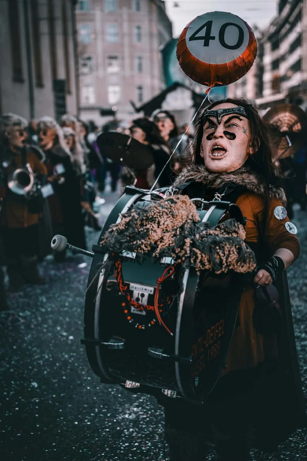 Eine Person mit schwarzen Gesichtsbalis, Brille und Kapuze auf einem Menschenauflauf beim Karneval. Sie fährt auf einem Fahrrad, das mit Dekorationen versehen ist, und hält einen roten Ballon mit der Zahl 40.
