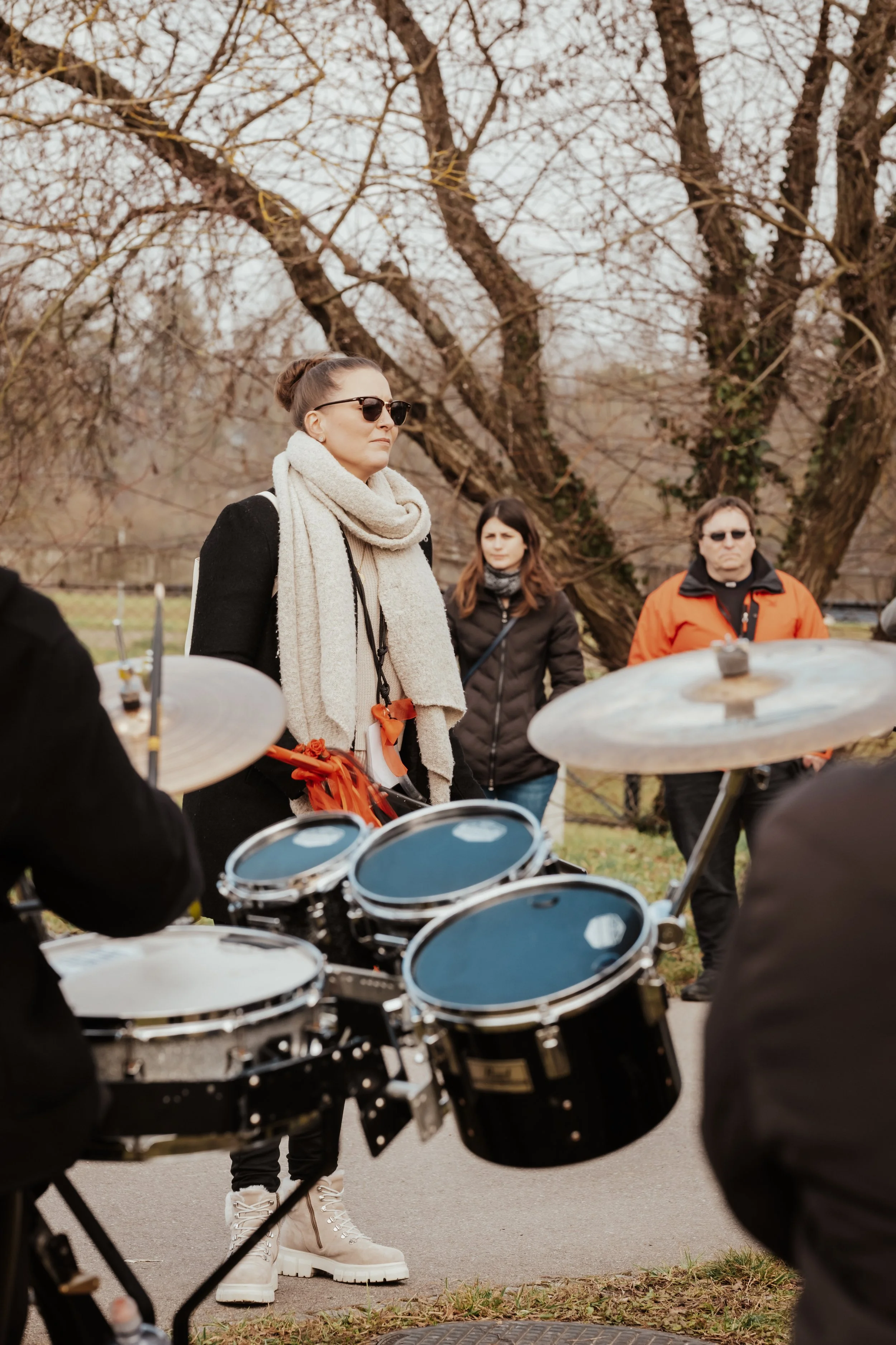 Eine Gruppe von Menschen, darunter eine Frau mit Sonnenbrille, Schal und dunklem Mantel, die anscheinend einer Musikprobe im Freien bei kaltem Wetter zuhört. Im Vordergrund sind Teil eines Schlagzeugs zu sehen, im Hintergrund Bäume ohne Blätter.