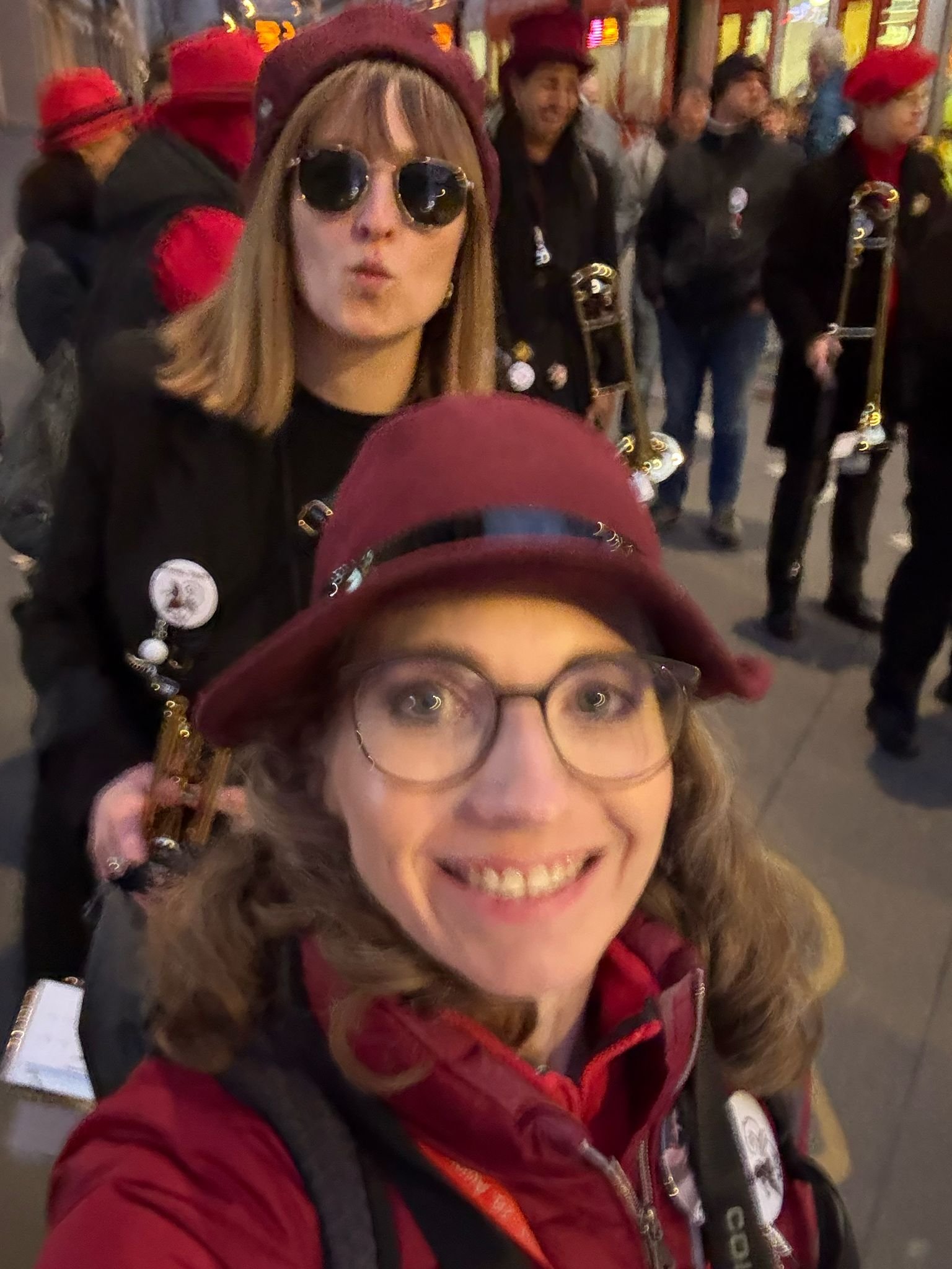 Zwei Frauen in roter Kleidung und Hut bei einer Parade oder Veranstaltung, im Hintergrund weitere Teilnehmer mit Musikinstrumenten.