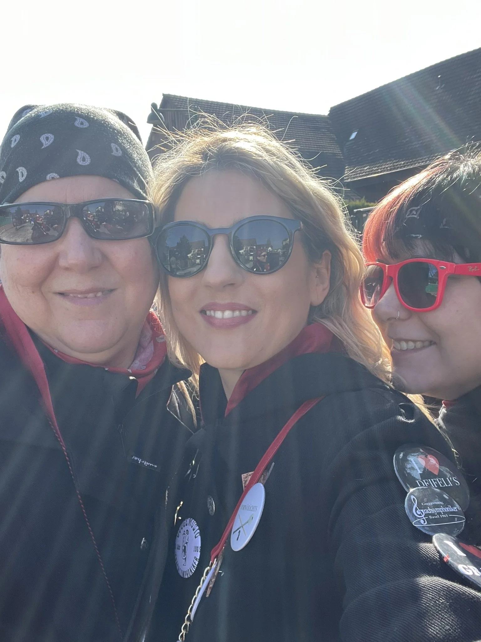 Drei Frauen tragen Sonnenbrillen und schwarze Jacken, einige mit Abzeichen und Pins, vor einem Sonnenhimmel und Häusern im Hintergrund.