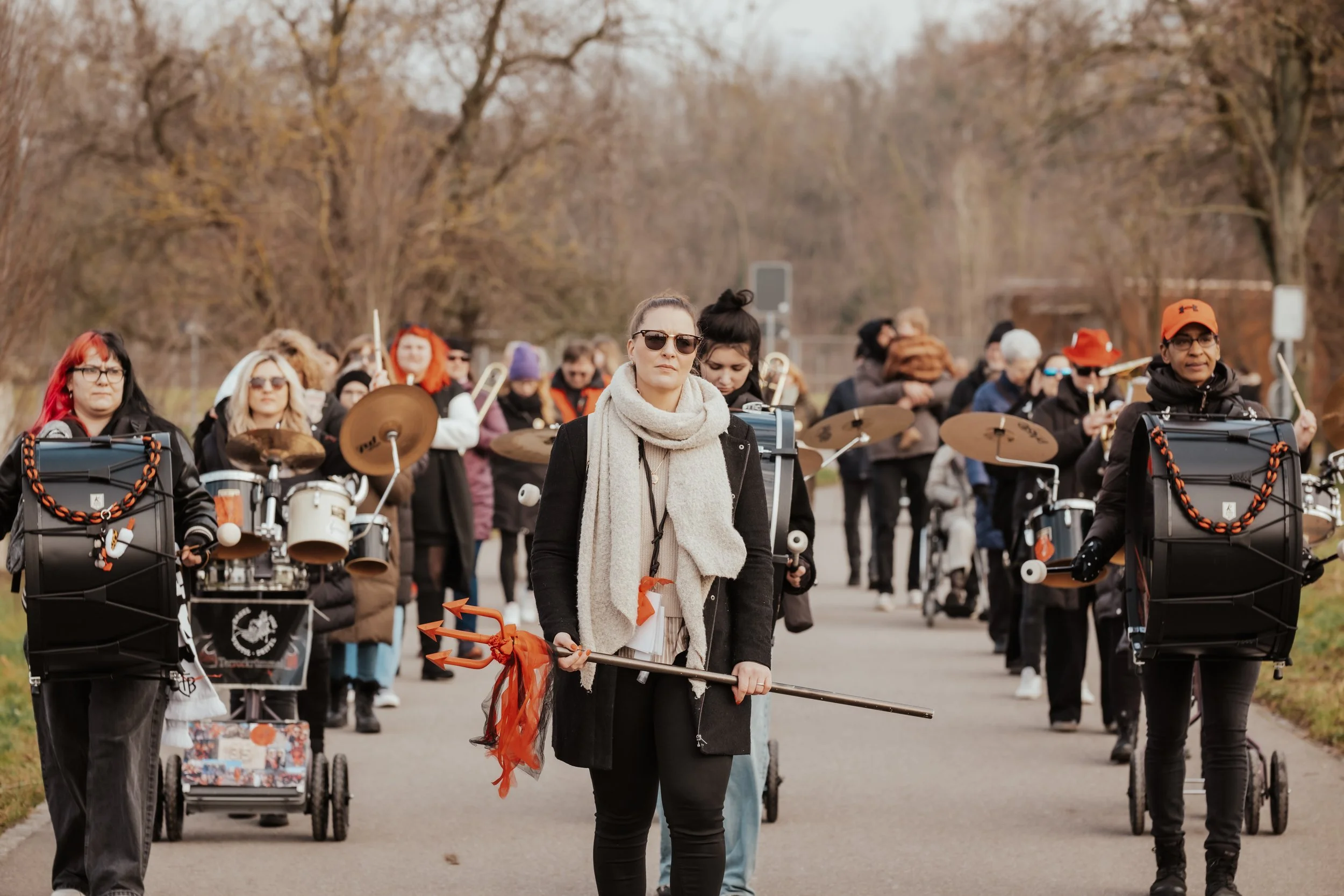 Eine Gruppe von Menschen, die bei einer Demonstration oder Parade im Freien marschieren, einige tragen Musikinstrumente, andere halten Banner oder Fahnen, im Hintergrund sind Bäume ohne Blätter.