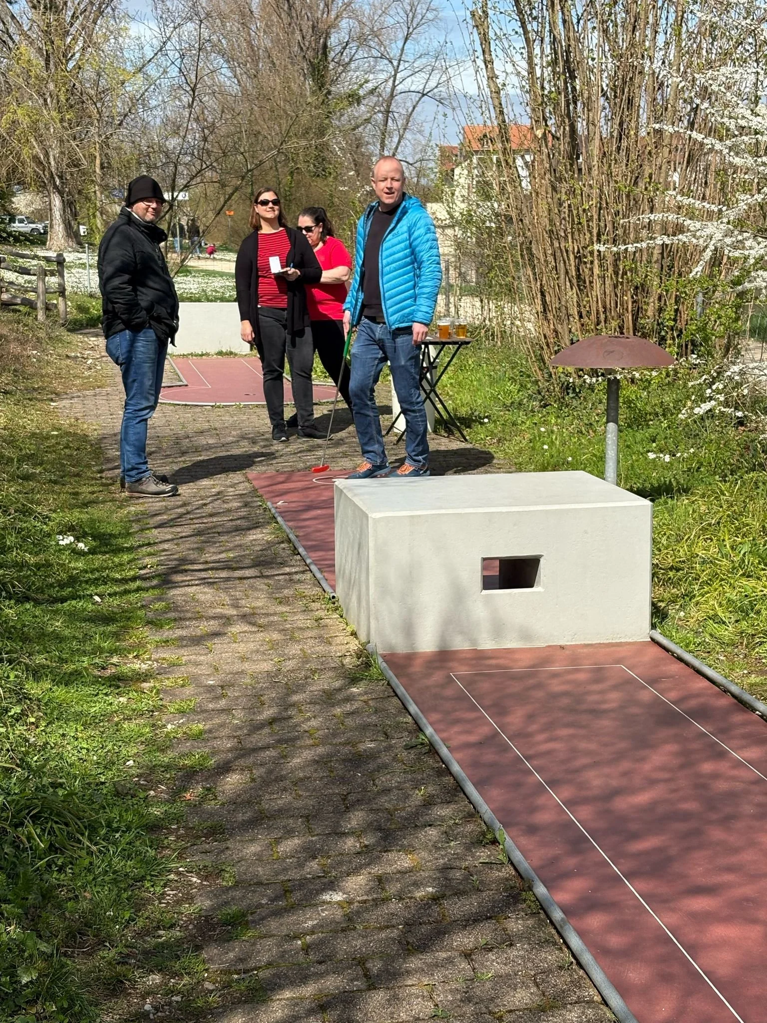 Vier Menschen stehen auf einer Minigolfanlage im Freien, eine Person bereitet den Abschlag vor, während die anderen zuschauen. Im Hintergrund sind Bäume und ein Gebäude sichtbar.
