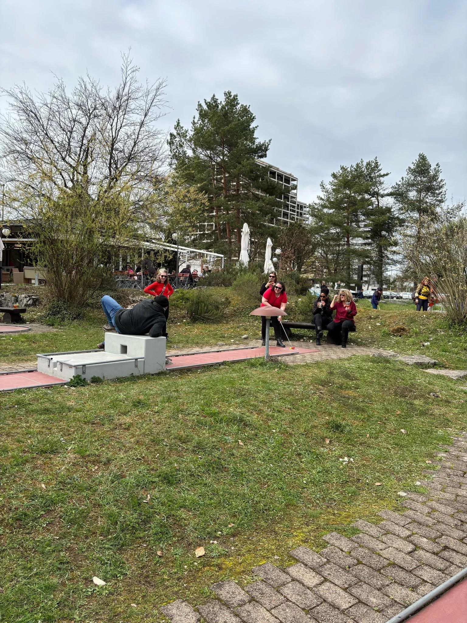 Menschen auf einer Garten- oder Parkfläche im Gespräch, einige sitzen auf Bänken, andere stehen, bei bewölktem Himmel.